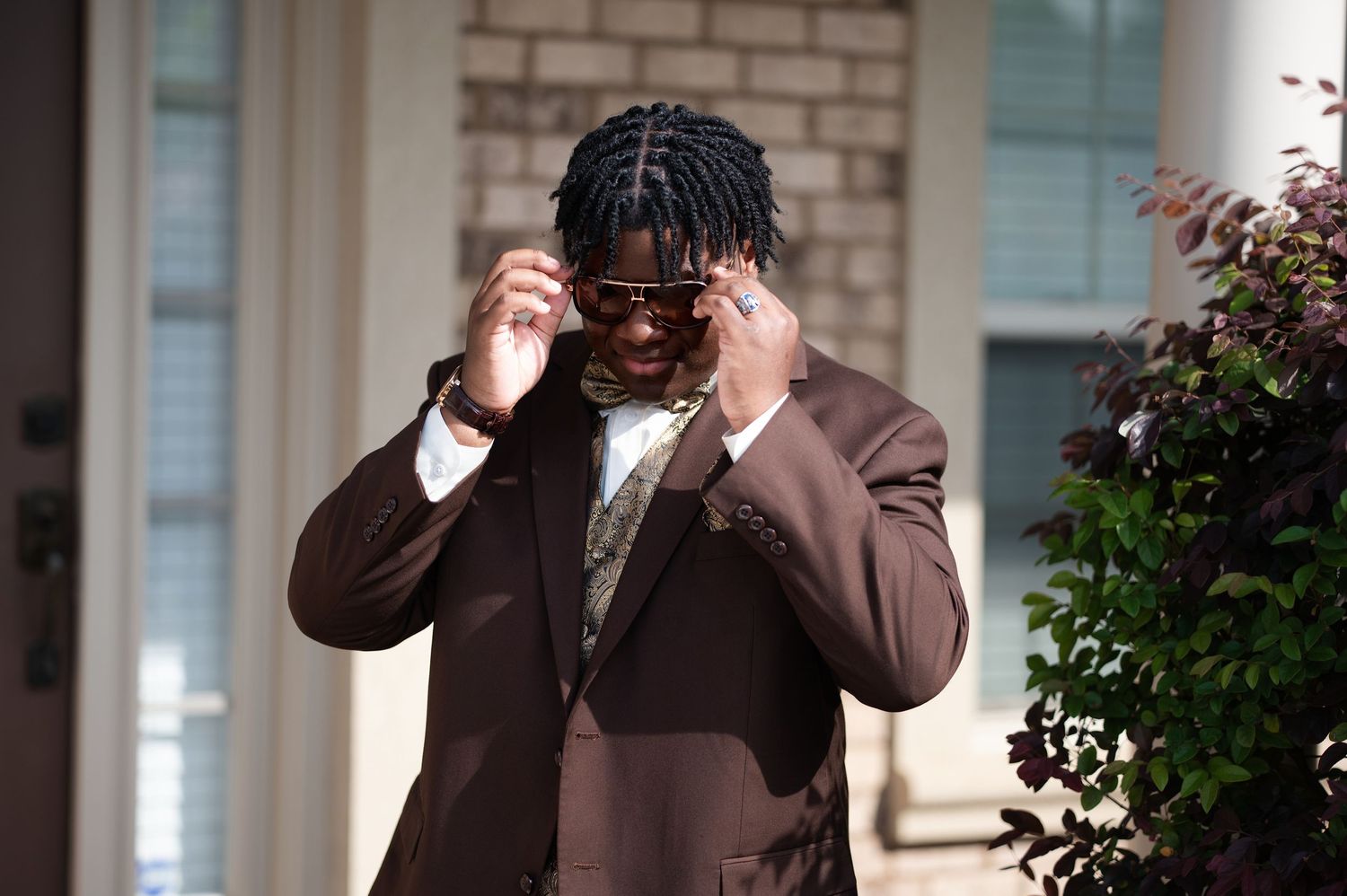 Stylish man in brown suit adjusting sunglasses outdoors, wearing bow tie and pocket square.