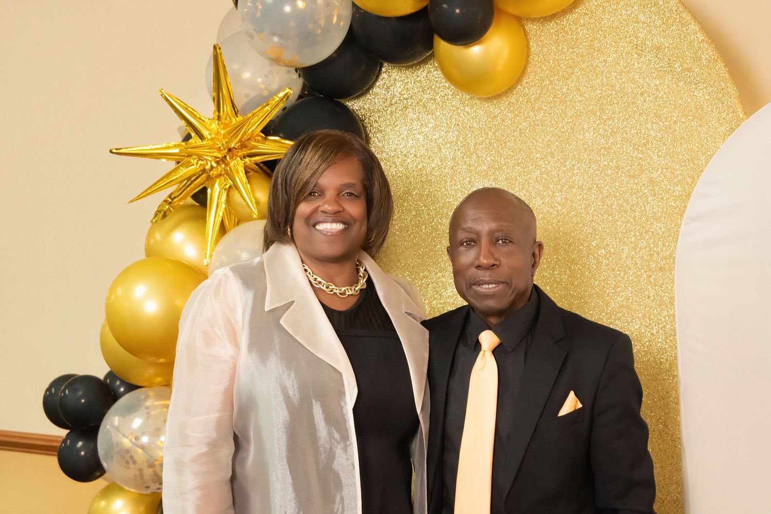 Two people smiling together in front of a gold glitter backdrop with black and gold balloon arch decorations.