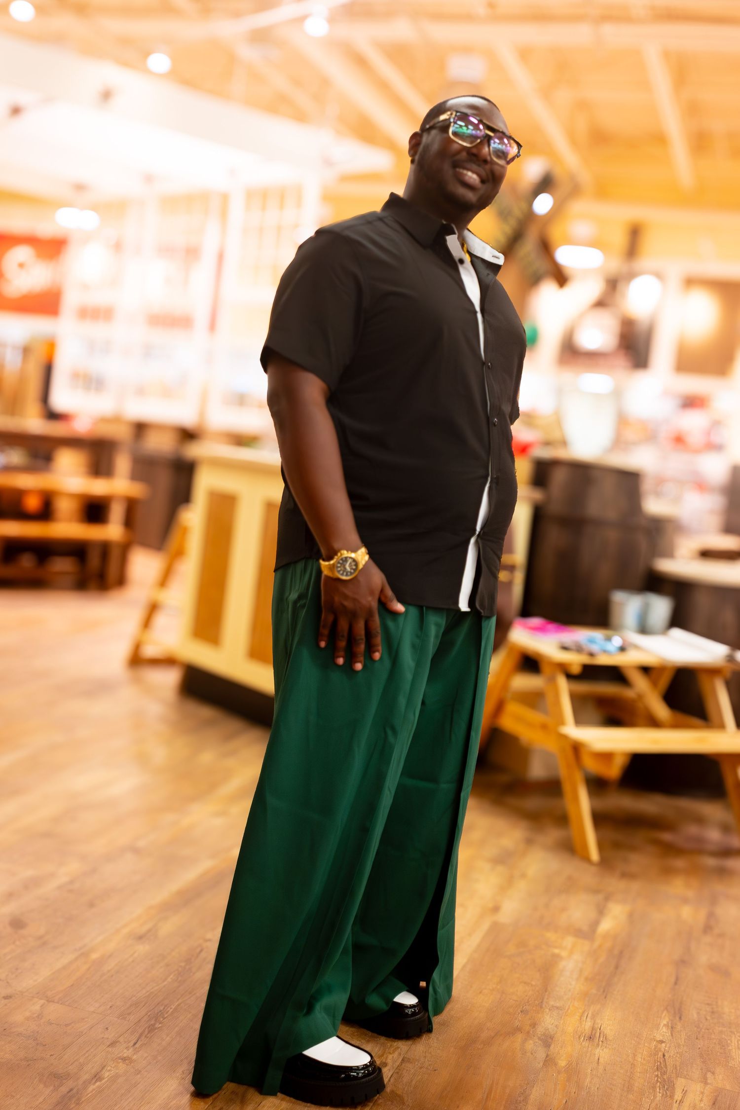 A person wearing a black shirt and green pants stands on a wooden floor in a warmly lit indoor space.