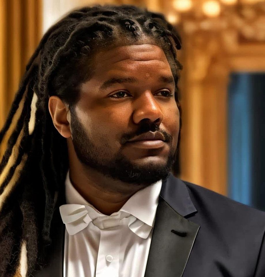 A young Black man with dreadlocks wearing a formal black suit and white dress shirt in an elegant setting.
