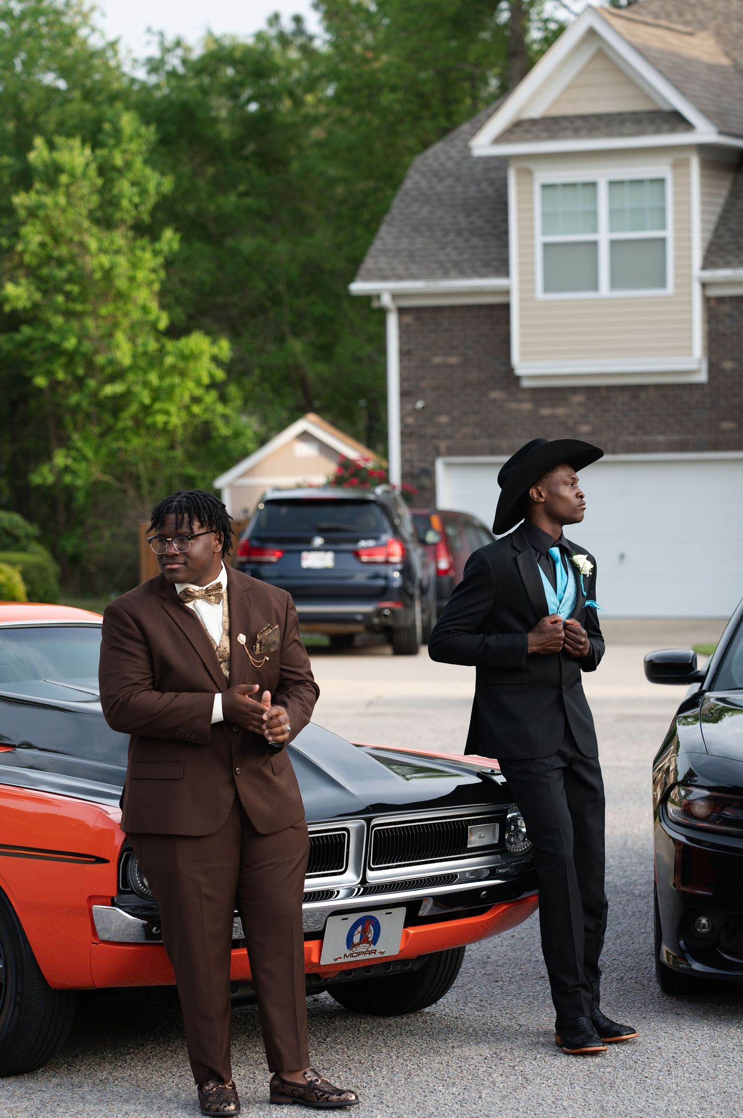 Two young men in formal suits pose beside a classic orange muscle car in a residential driveway.