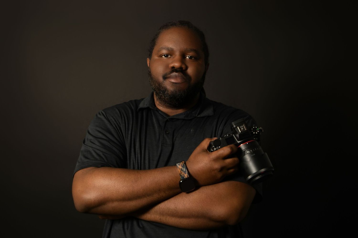 A professional male photographer poses with arms crossed holding a camera, wearing a black shirt against a dark background.