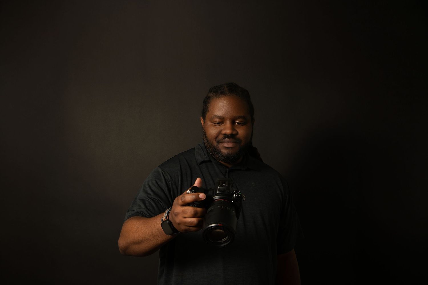 A man in a black shirt holds a professional DSLR camera against a dark studio background.