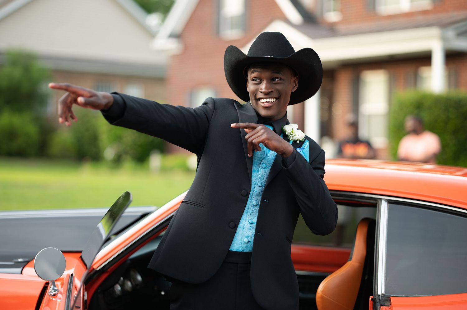 Young man in black suit and cowboy hat pointing while leaning on an orange car at a prom event.
