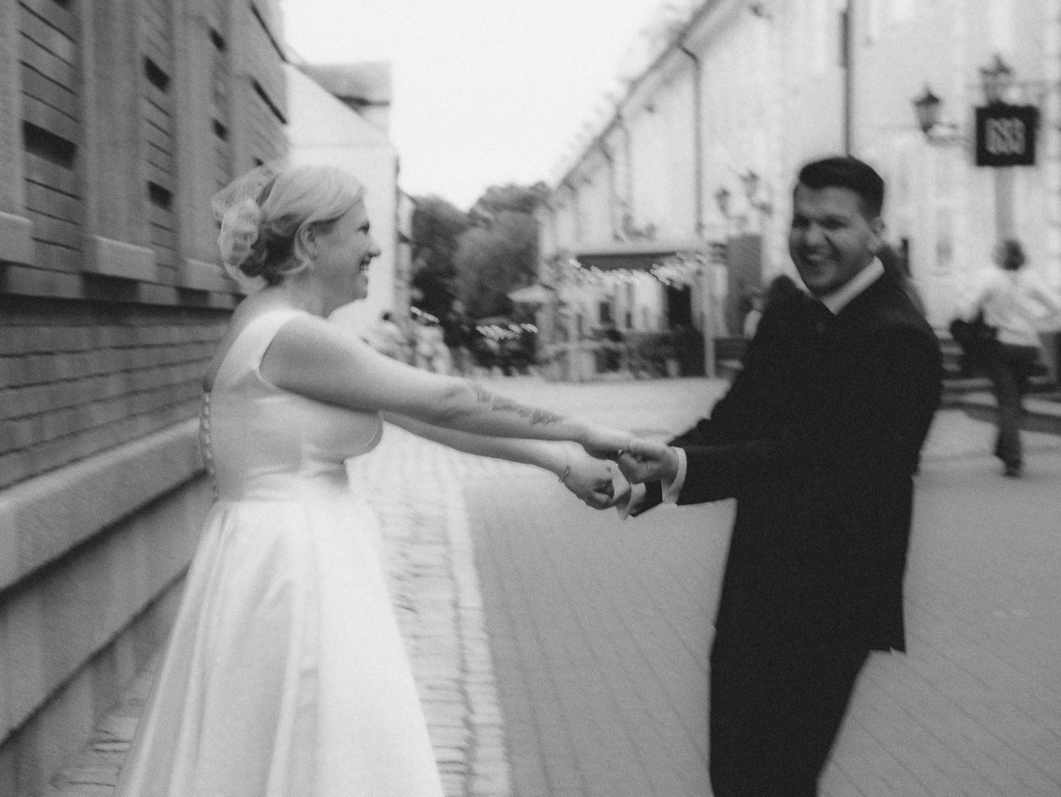 Bride and groom holding hands and dancing playfully on a cobblestone street in black and white photo.