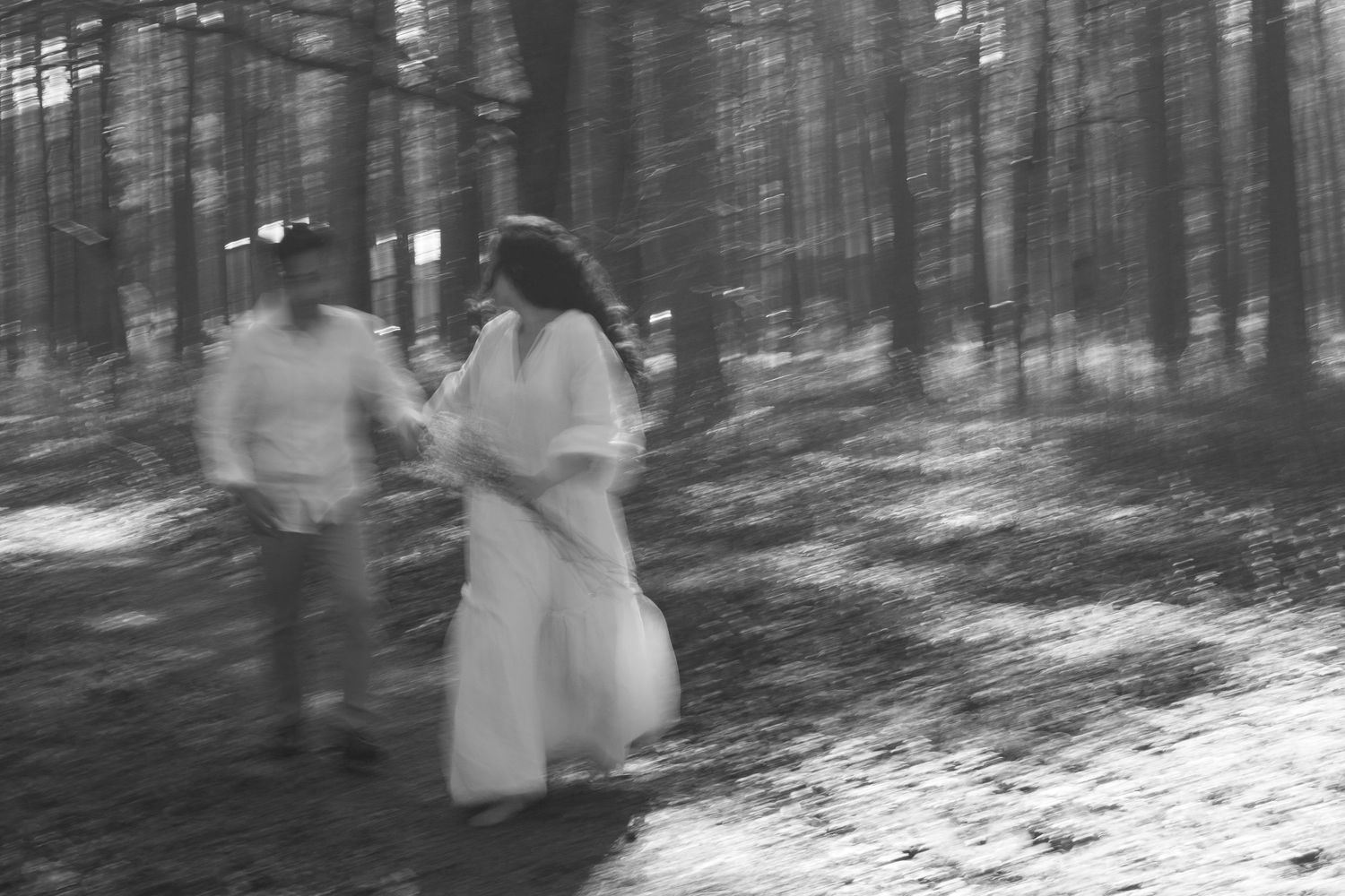 Blurred black and white photo of couple running through pine forest in white outfits.