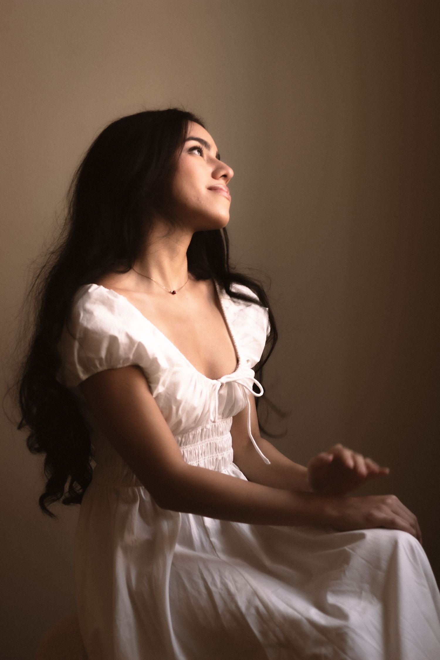 Young woman in white dress sitting and looking upward in dramatic side lighting against dark background.