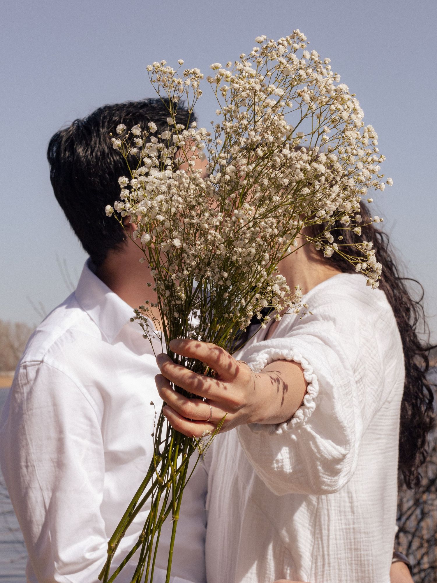Couple sharing kiss hidden behind large bouquet of white wildflowers by riverside.