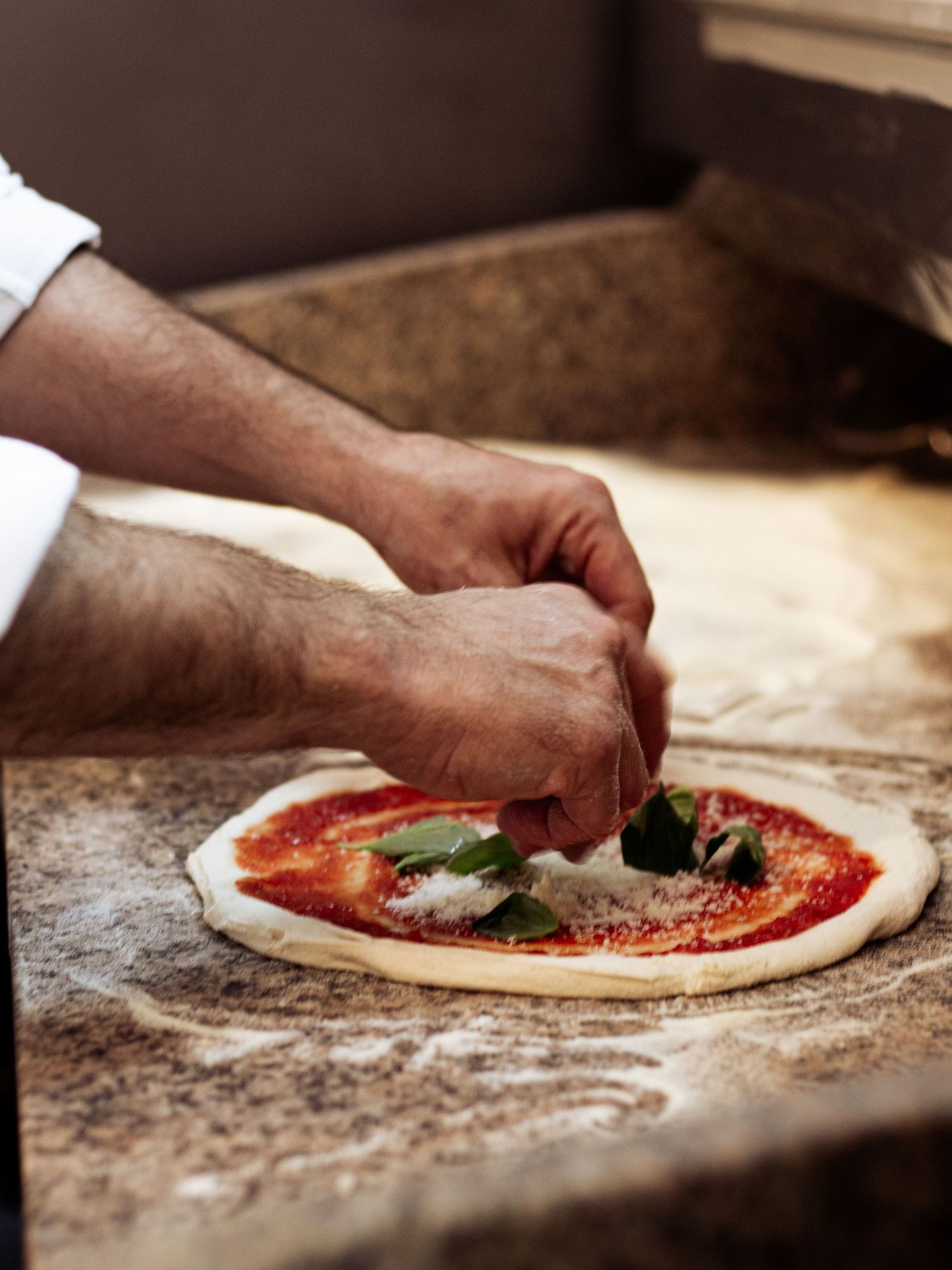 Chef hands placing fresh basil leaves on a raw Neapolitan pizza with tomato sauce and mozzarella.
