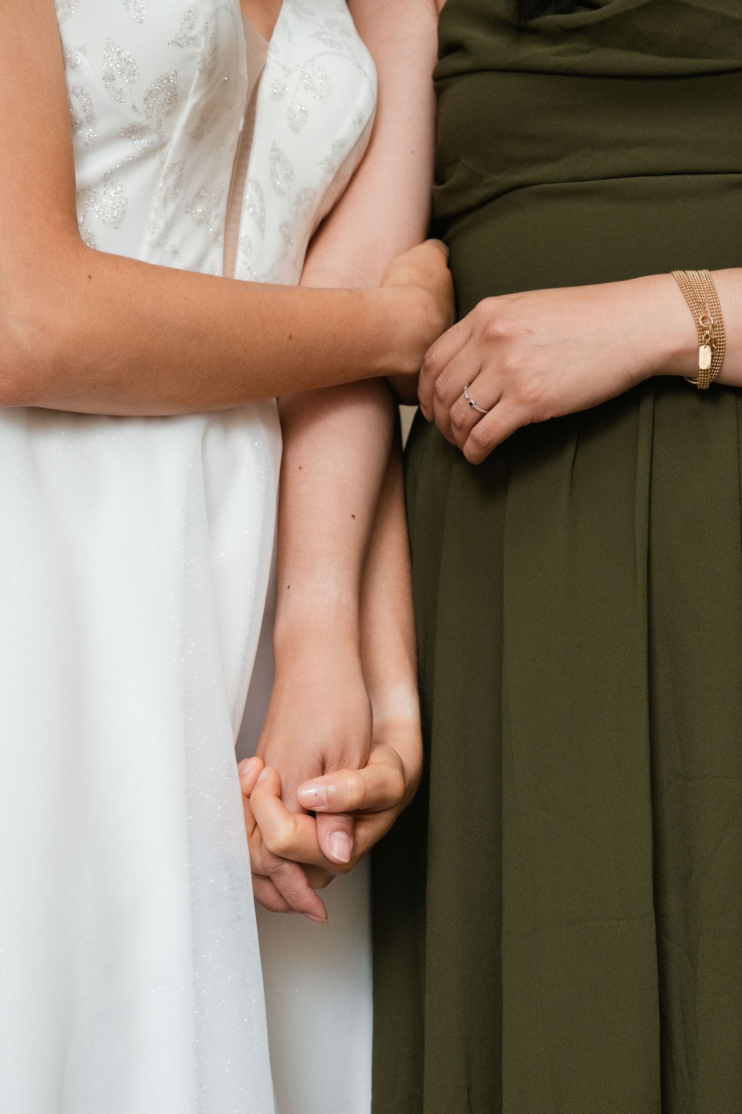 Two women in elegant dresses hold hands, one in white bridal gown and one in olive green, wearing gold jewelry.