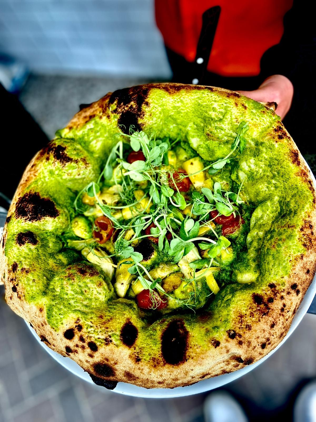 Vibrant green pesto Neapolitan pizza topped with avocado, tomatoes, and microgreens.