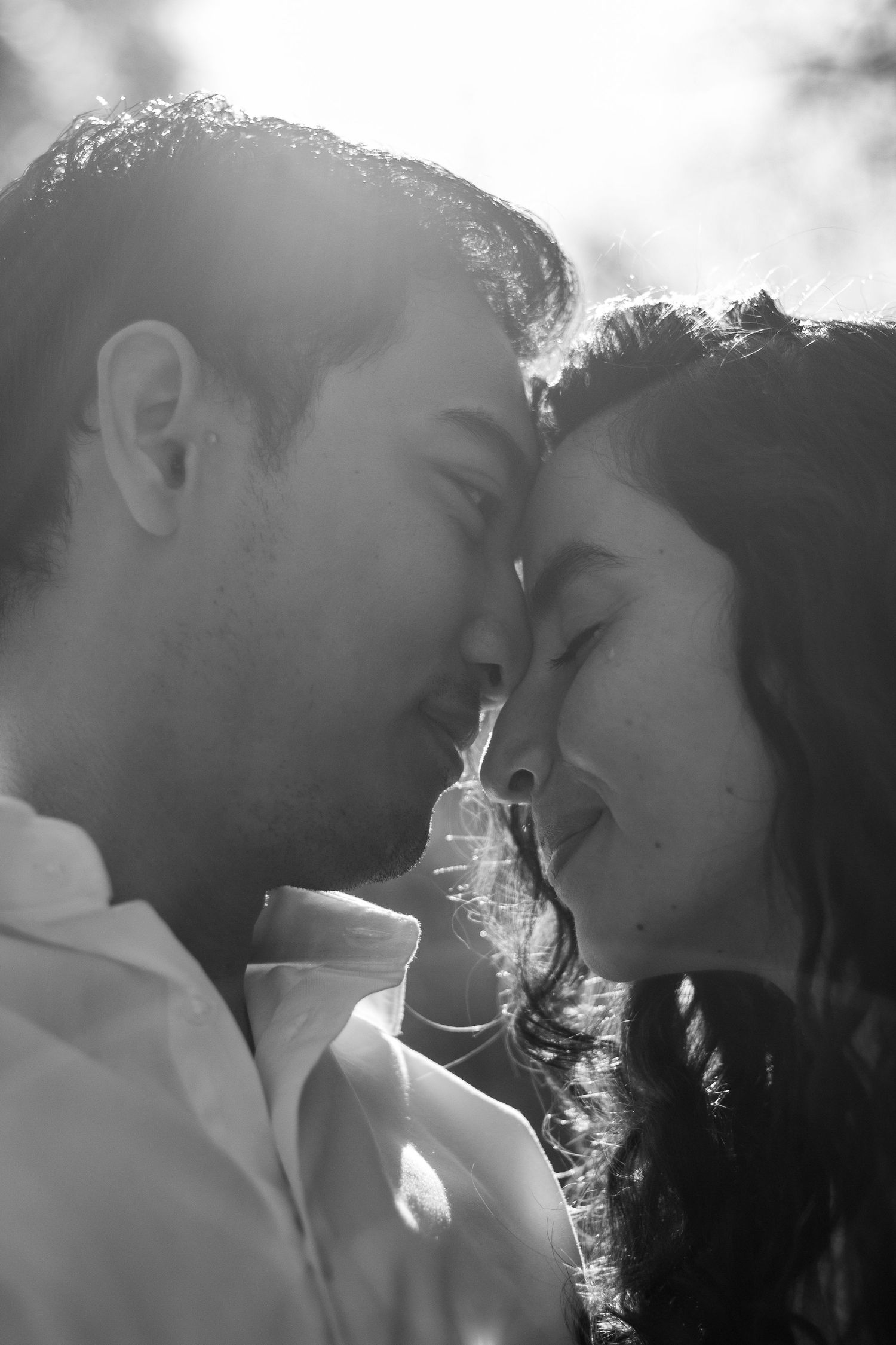 Romantic black and white close-up of couple with foreheads touching, backlit by bright sunlight.