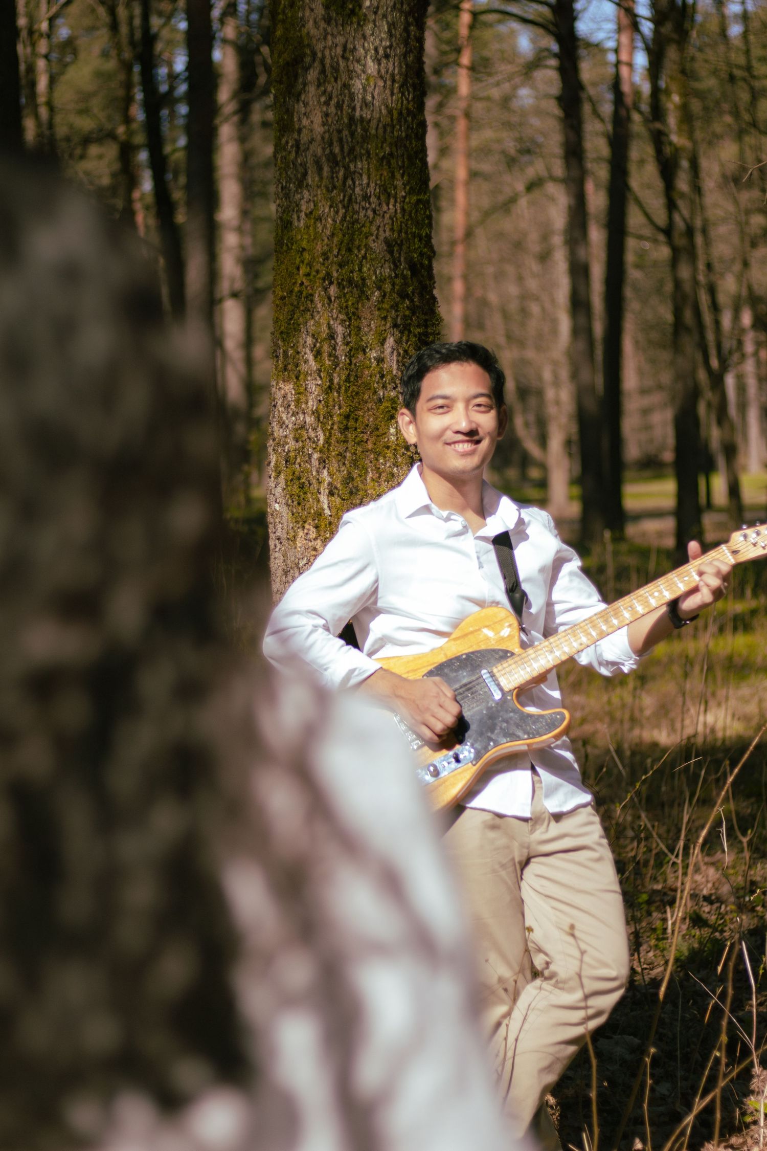 Smiling man playing electric guitar leaning against tree in sunny forest setting.