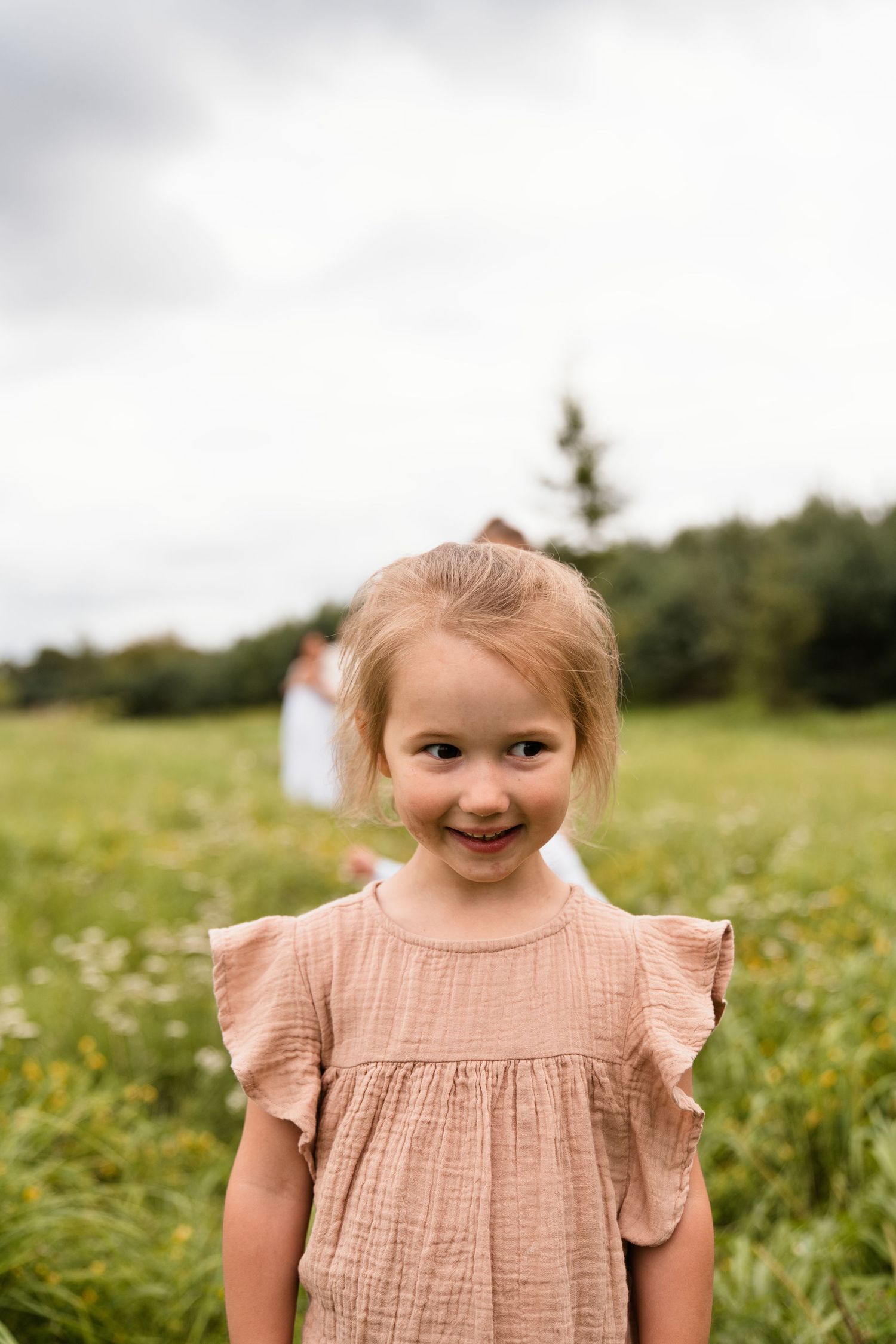 A smiling young girl in a pink ruffle dress stands in a green meadow with blurred figures and trees in the background.