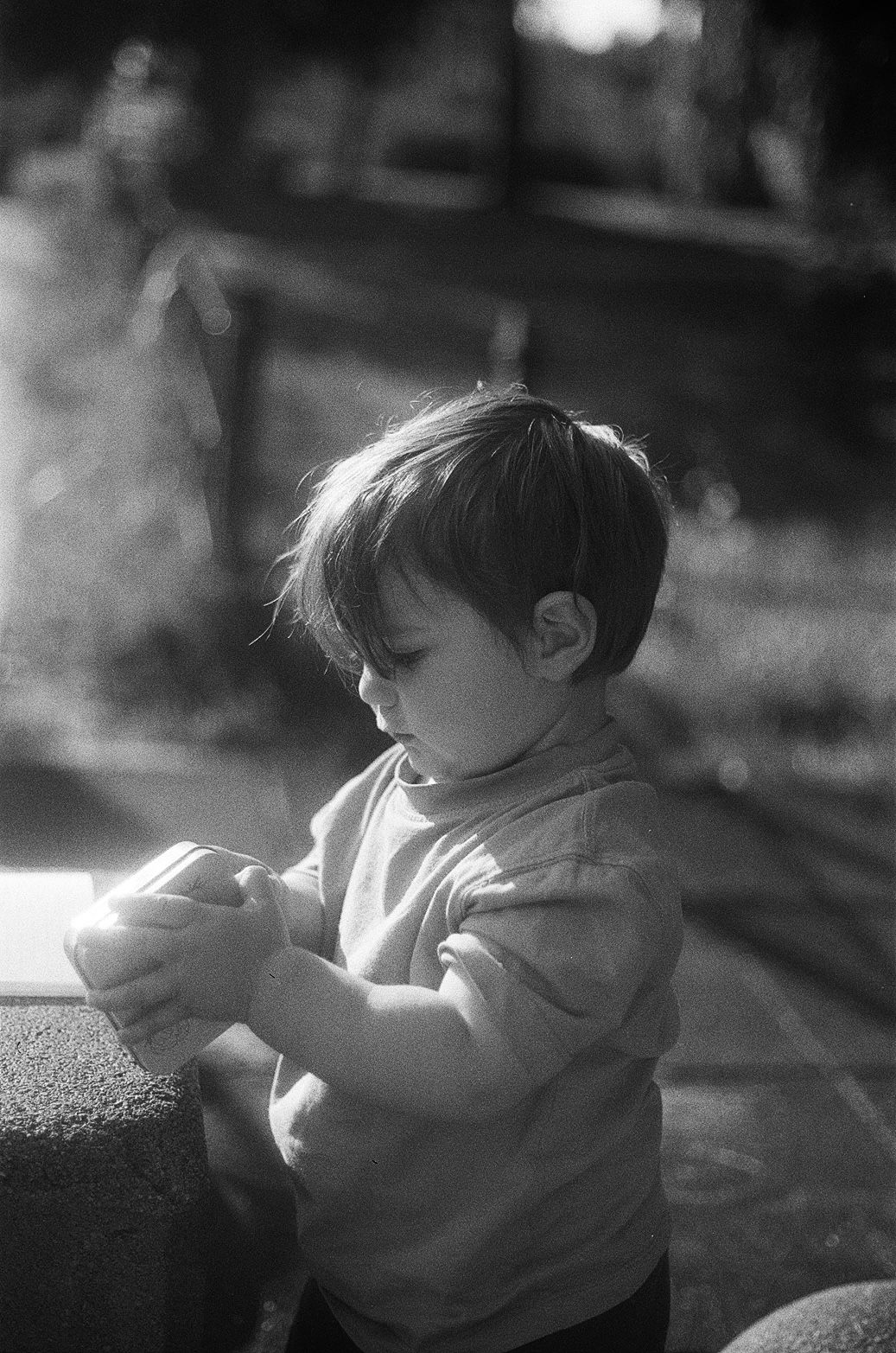 Black and white photo of a young toddler boy looking down at something in his hands outdoors.
