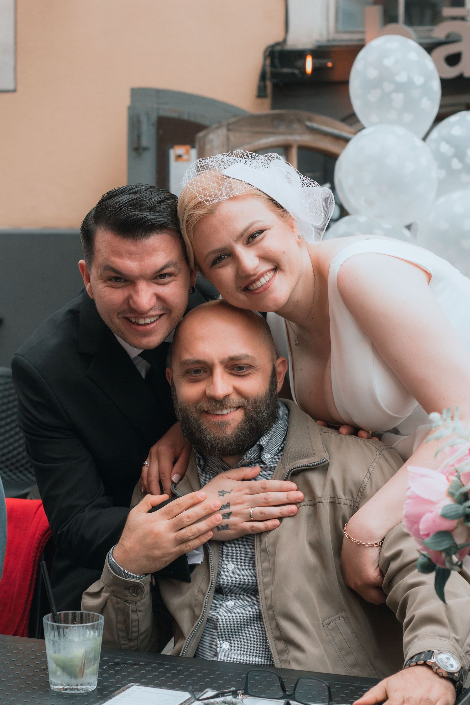 Bride in white veil and groom in black suit pose with bearded friend at wedding reception with balloons.