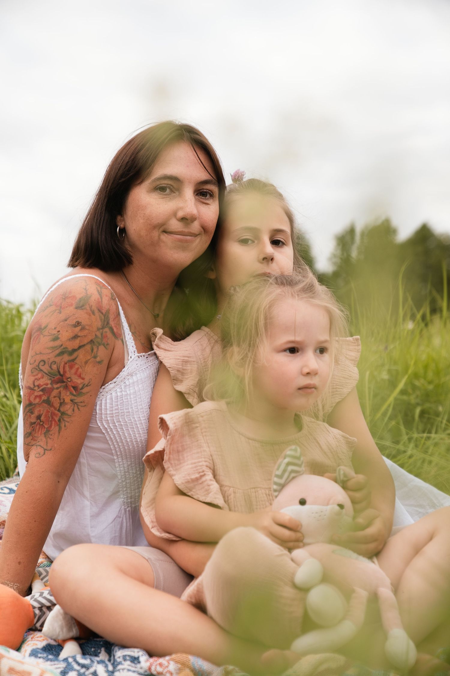 A tattooed mother sits outdoors on a blanket with her two young daughters, one holding a stuffed bunny toy.