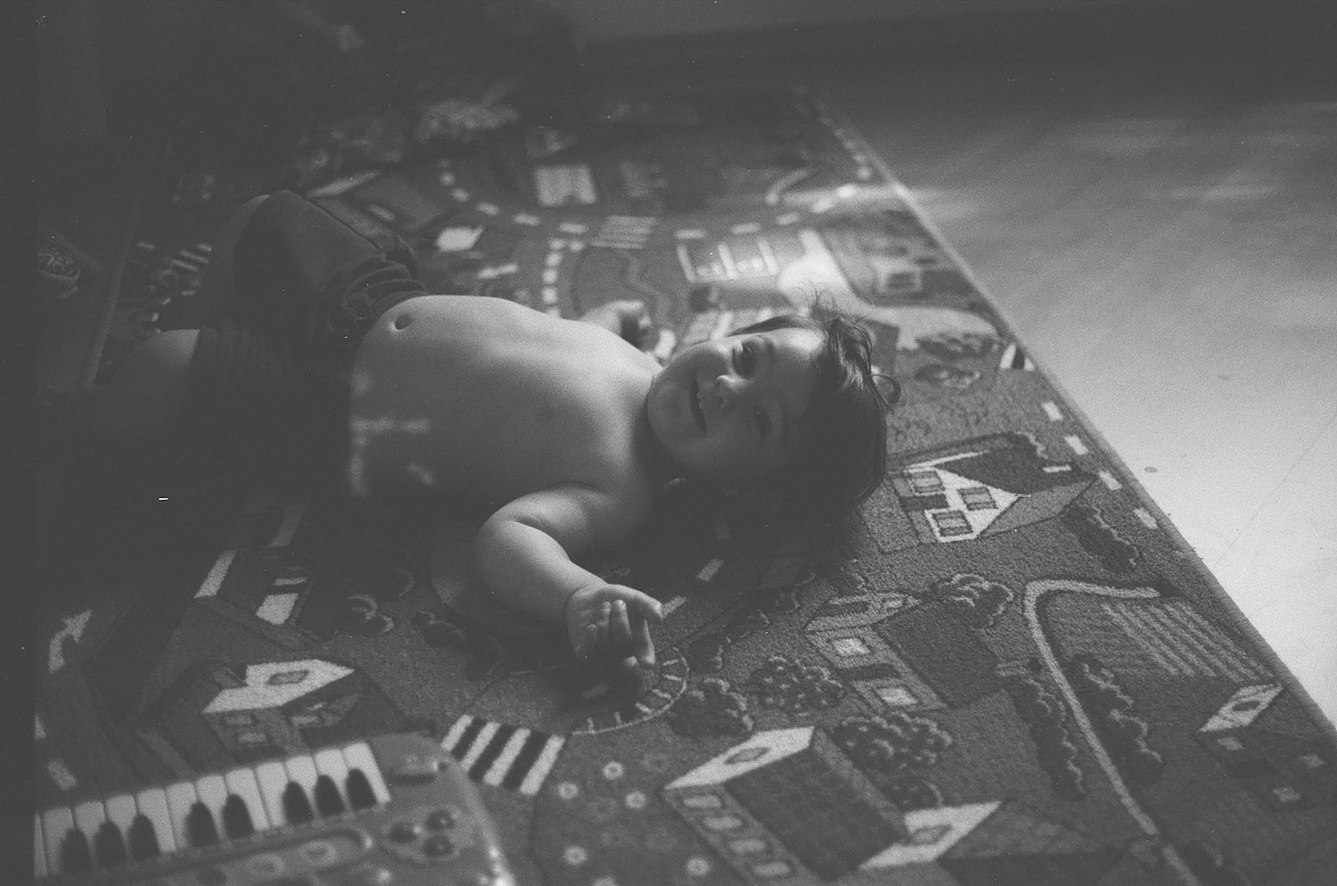 Black and white photo of a shirtless child lying on a colorful patterned play mat on the floor.