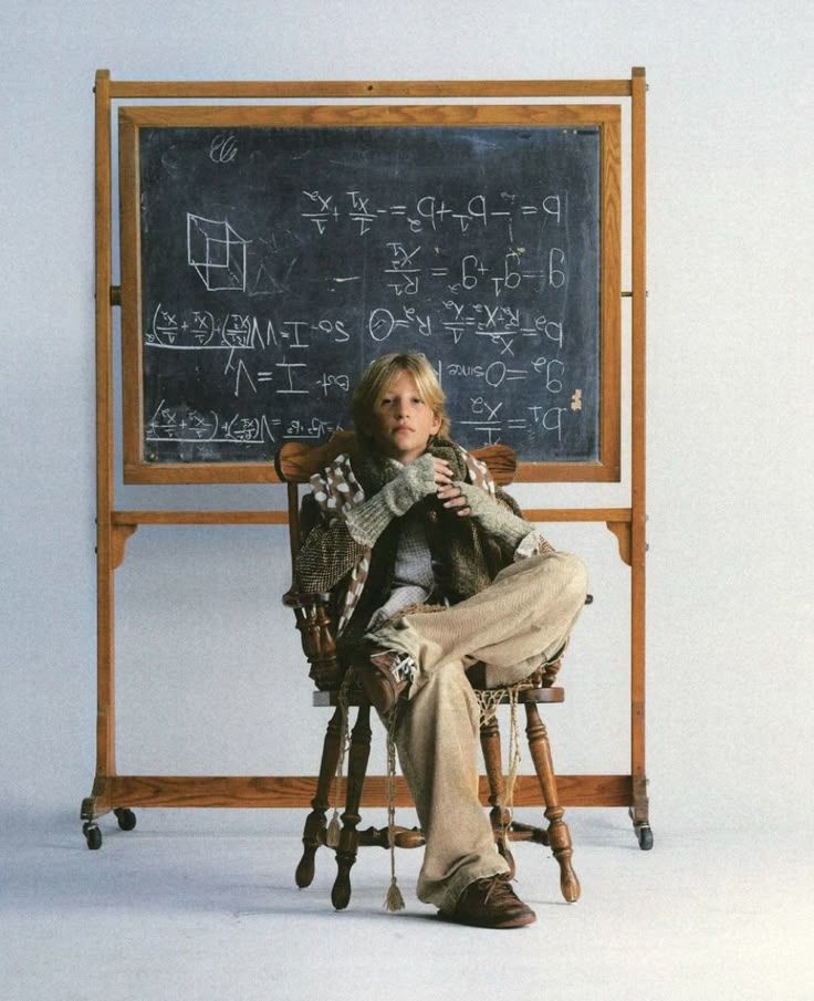 Young boy sitting in a wooden chair in front of a chalkboard filled with math equations.