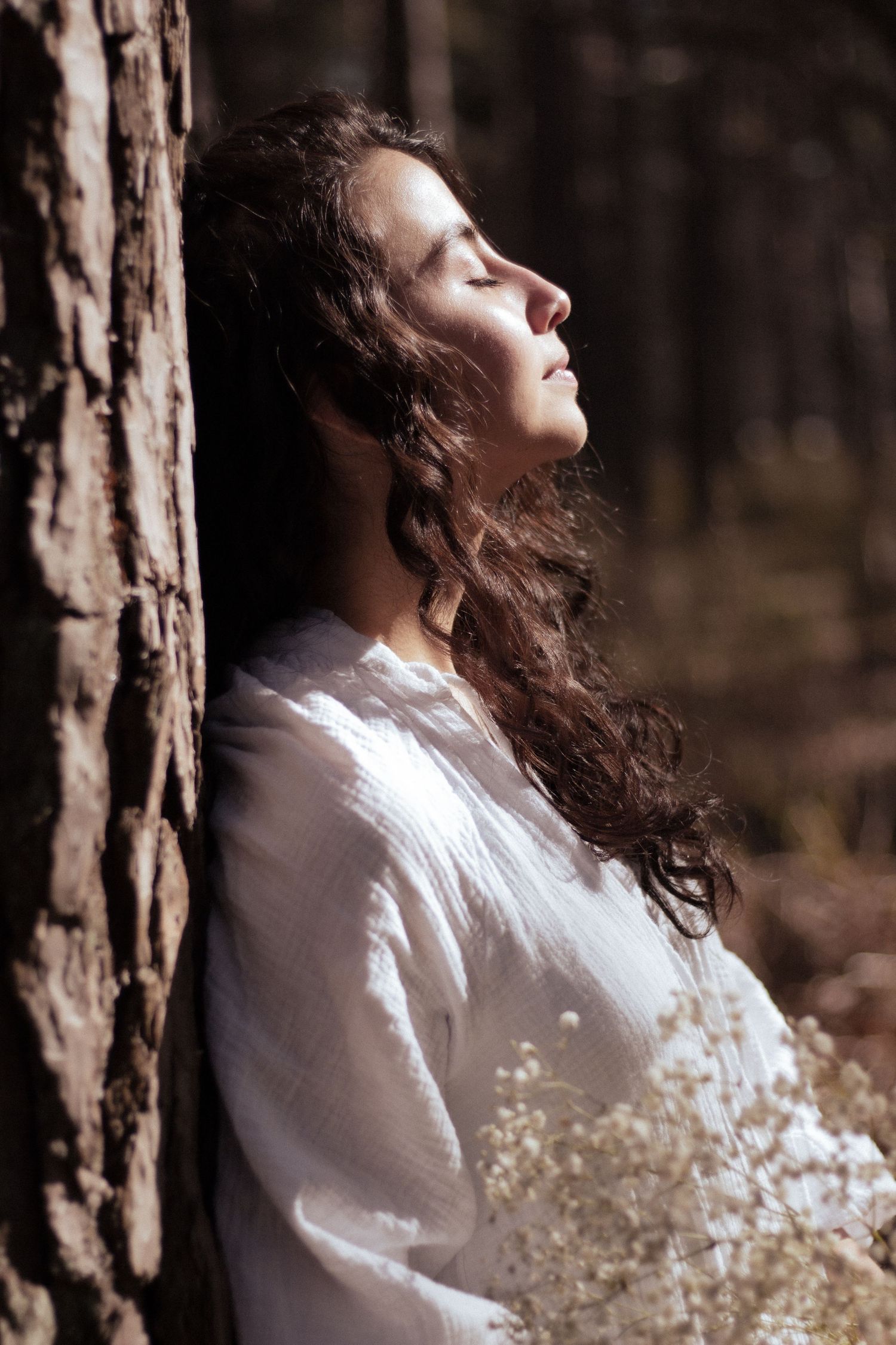 Woman in white linen dress leaning against pine tree in forest, eyes closed, enjoying sunlight.