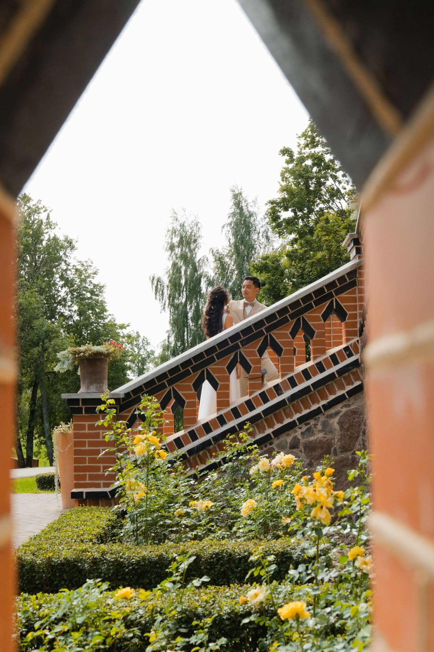 A couple embraces on decorative brick steps framed by yellow roses and hedges, viewed through an archway.