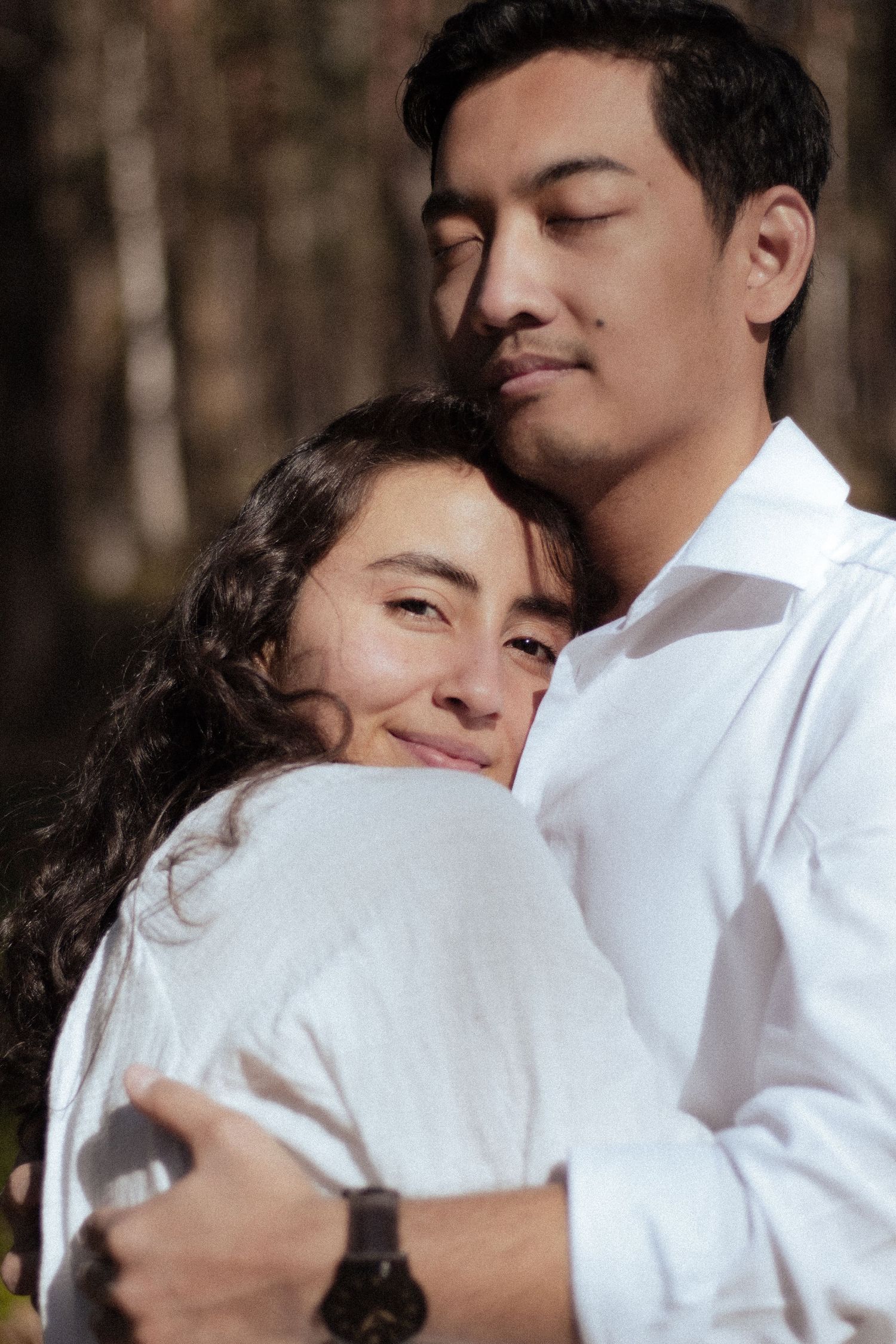 Tender moment of smiling woman resting head on mans chest as he embraces her in forest setting.