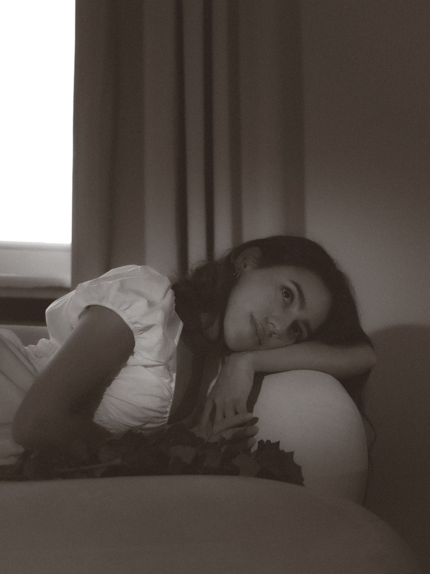 Black and white portrait of a young woman resting her head on her arms on a sofa near a window.