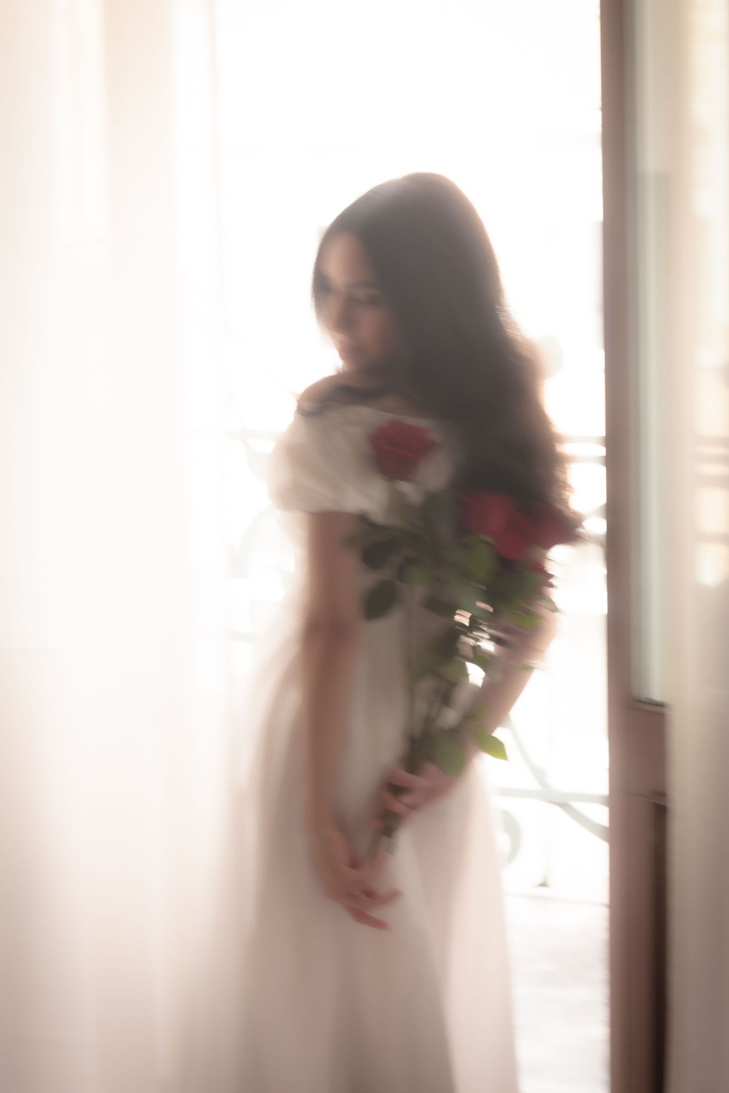 Dreamy blurred portrait of a young woman in a white off-shoulder dress holding red roses by a bright window.