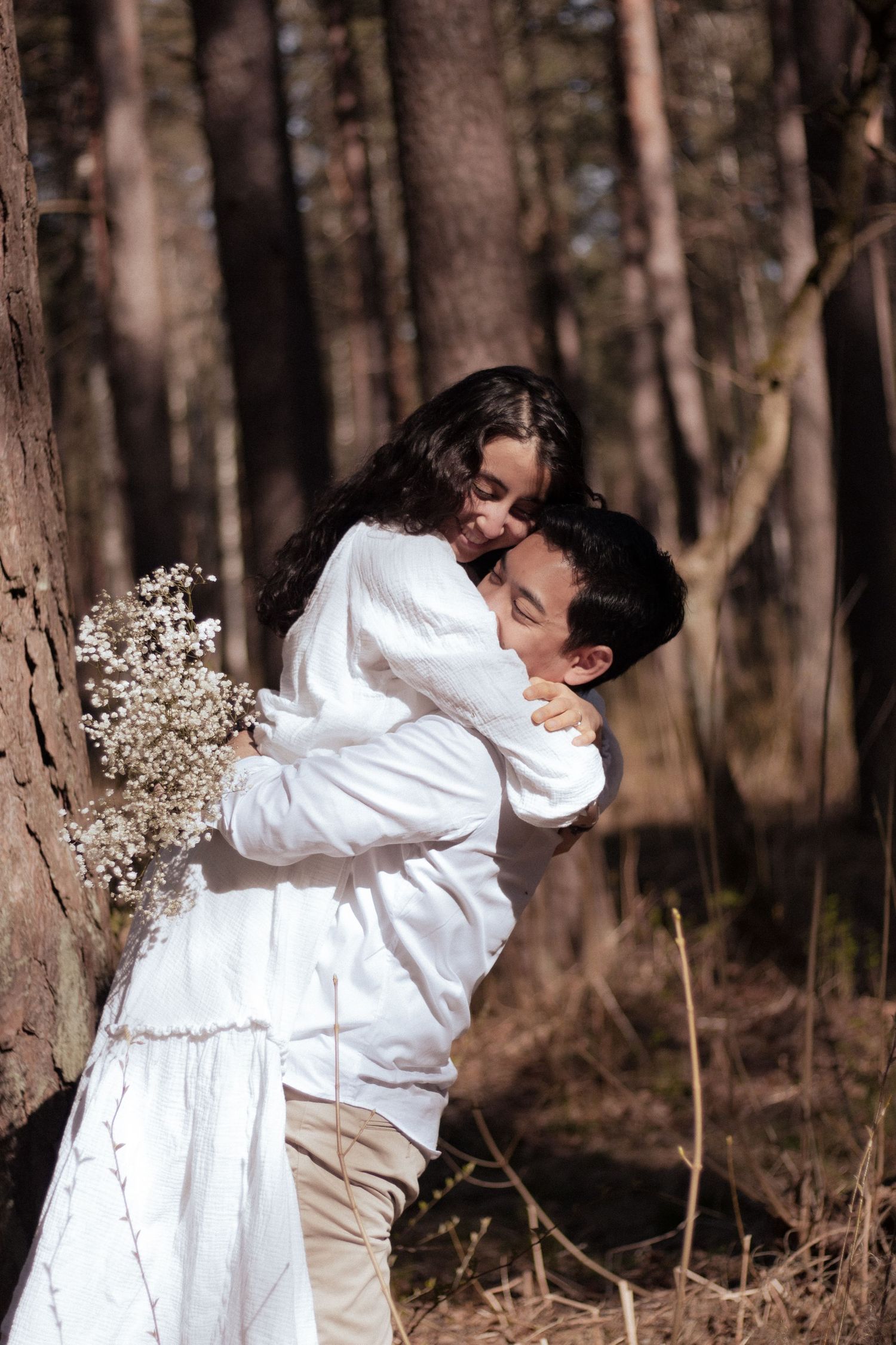 Romantic couple embracing in a sunlit forest, woman in white dress holding wildflowers, leaning against a tree.