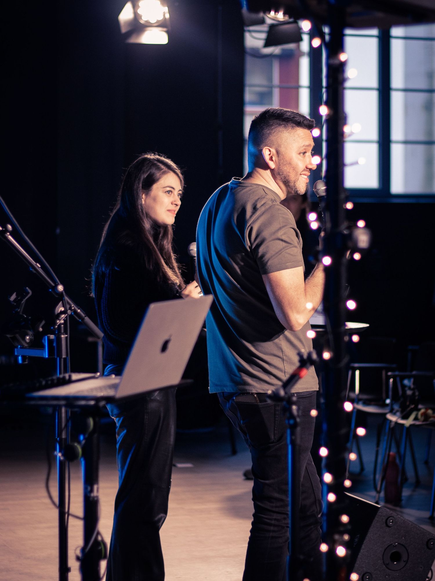 A woman and man standing on stage with a laptop and pixie lights behind them.