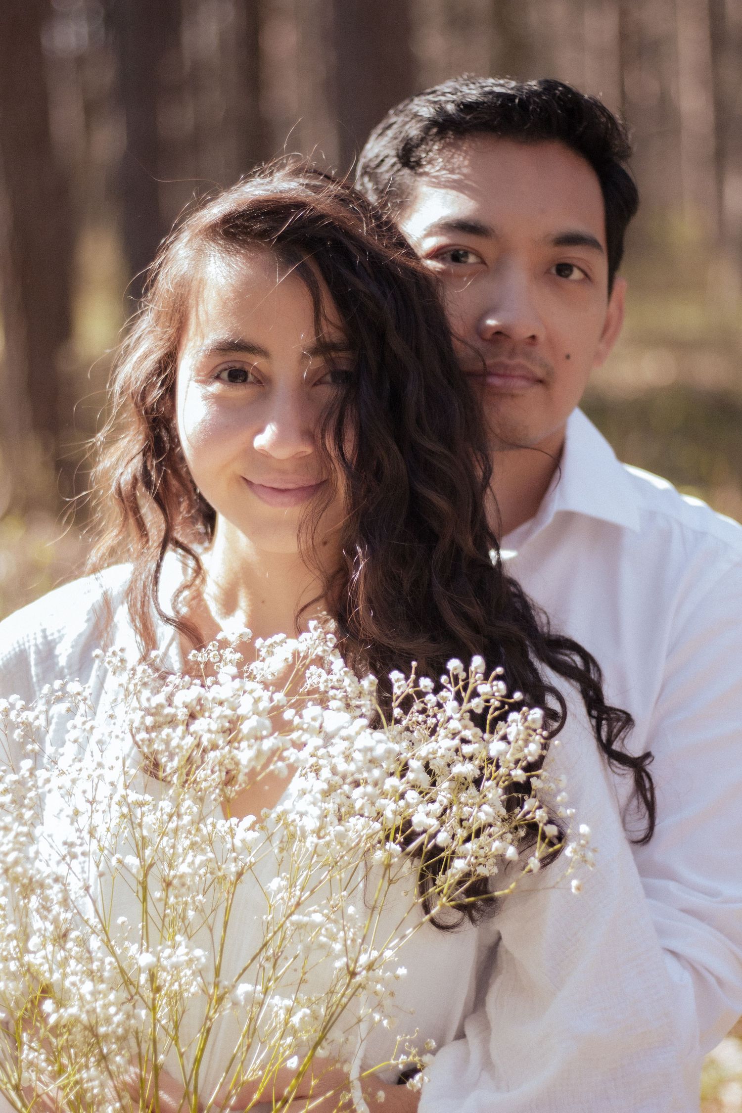Couple embracing in pine forest, woman holding white wildflowers bouquet, both wearing white outfits.