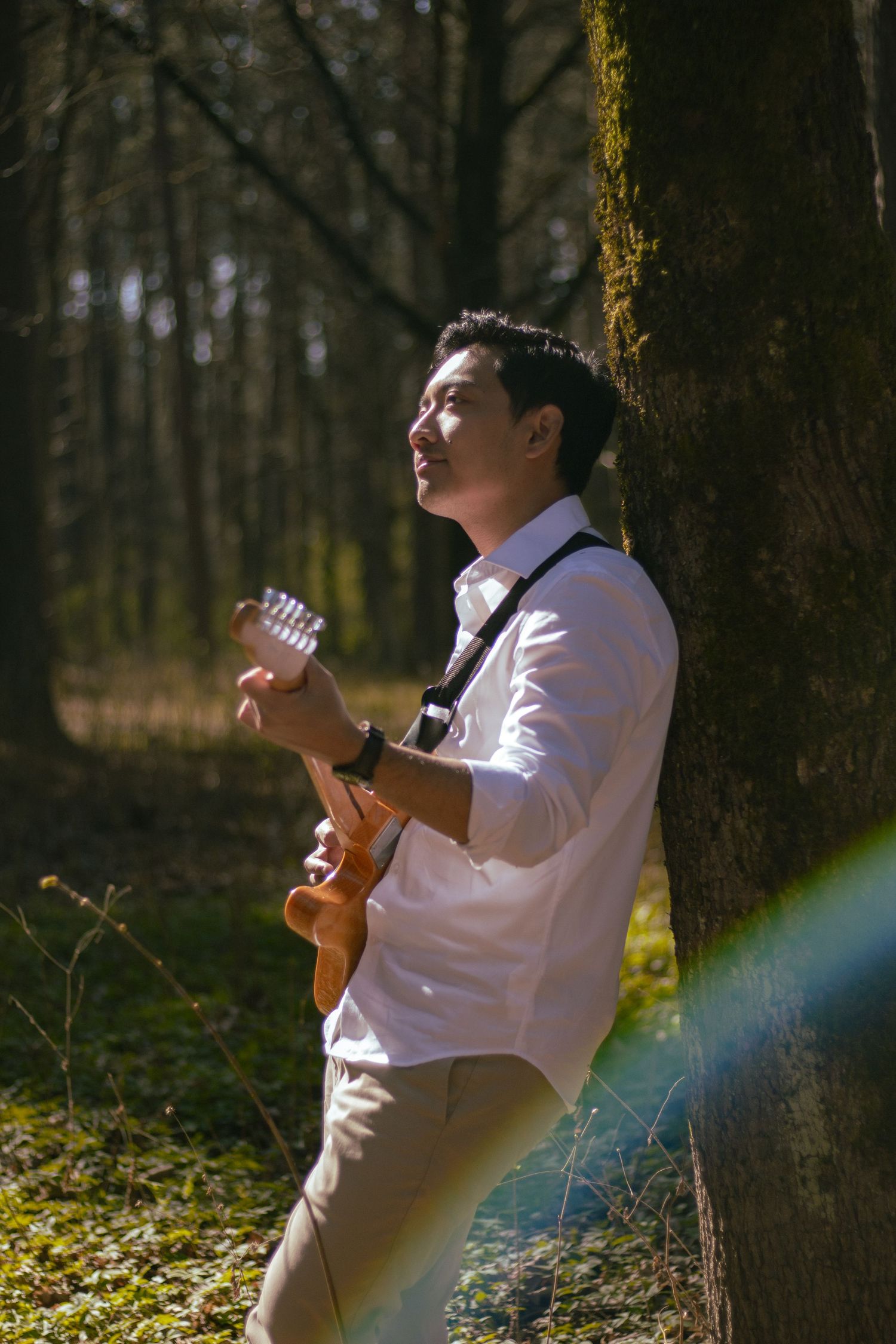 Man playing guitar leaning against moss-covered tree in sunlit forest with lens flare.