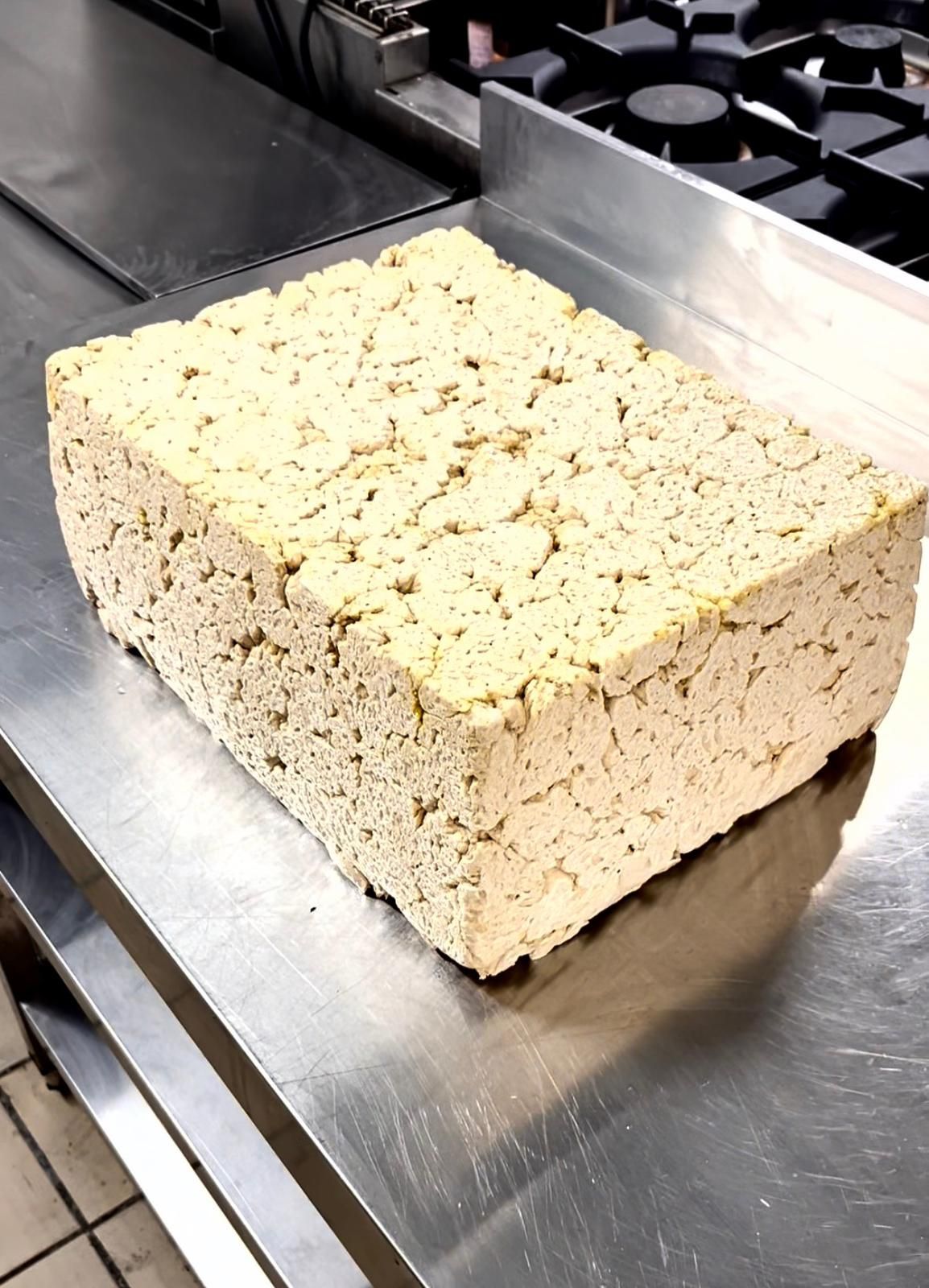 A large square block of firm tofu with a porous texture sitting on a stainless steel kitchen surface.