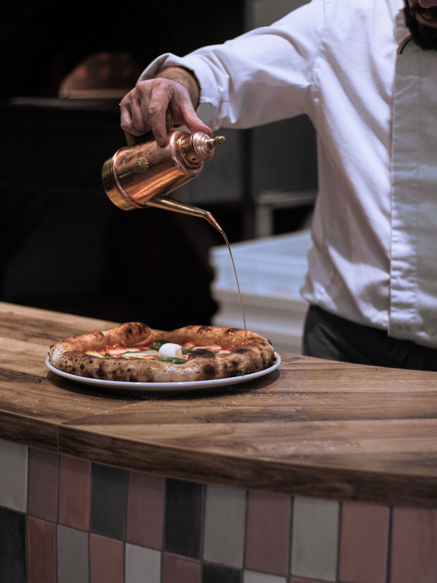 Chef drizzling olive oil from a copper bottle onto a freshly baked Neapolitan pizza on a rustic wooden counter.