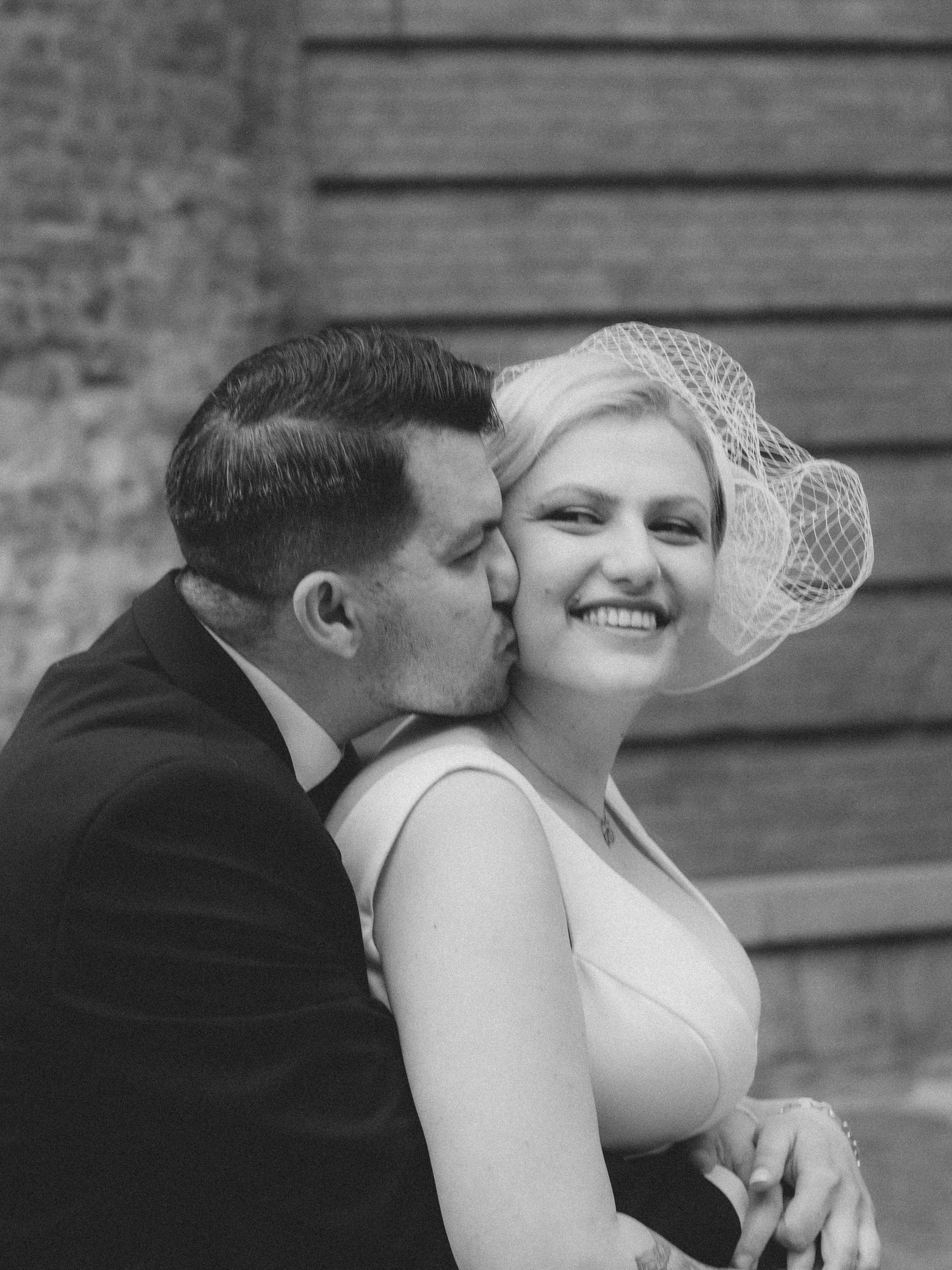 Black and white wedding photo of groom kissing bride on cheek, bride wearing birdcage veil and smiling.