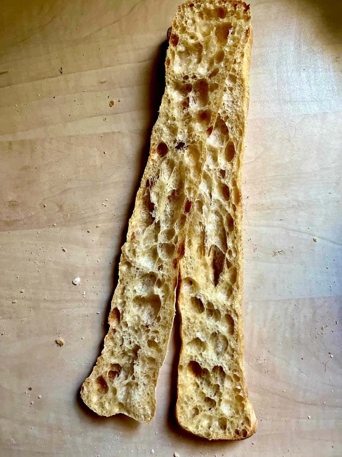 Long thin slice of artisan sourdough bread with large open holes and crispy crust on wooden surface.