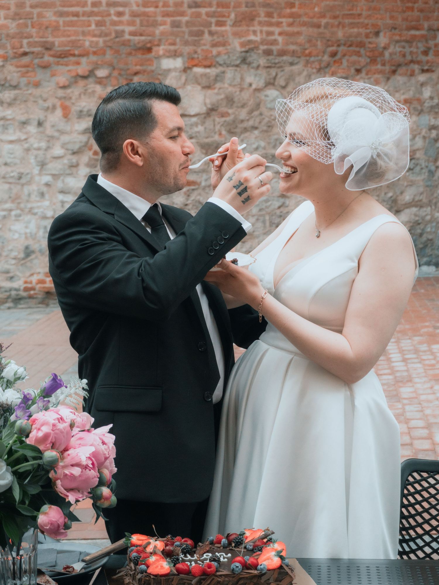 Bride and groom feeding each other wedding cake outdoors, with peonies and a brick wall backdrop.