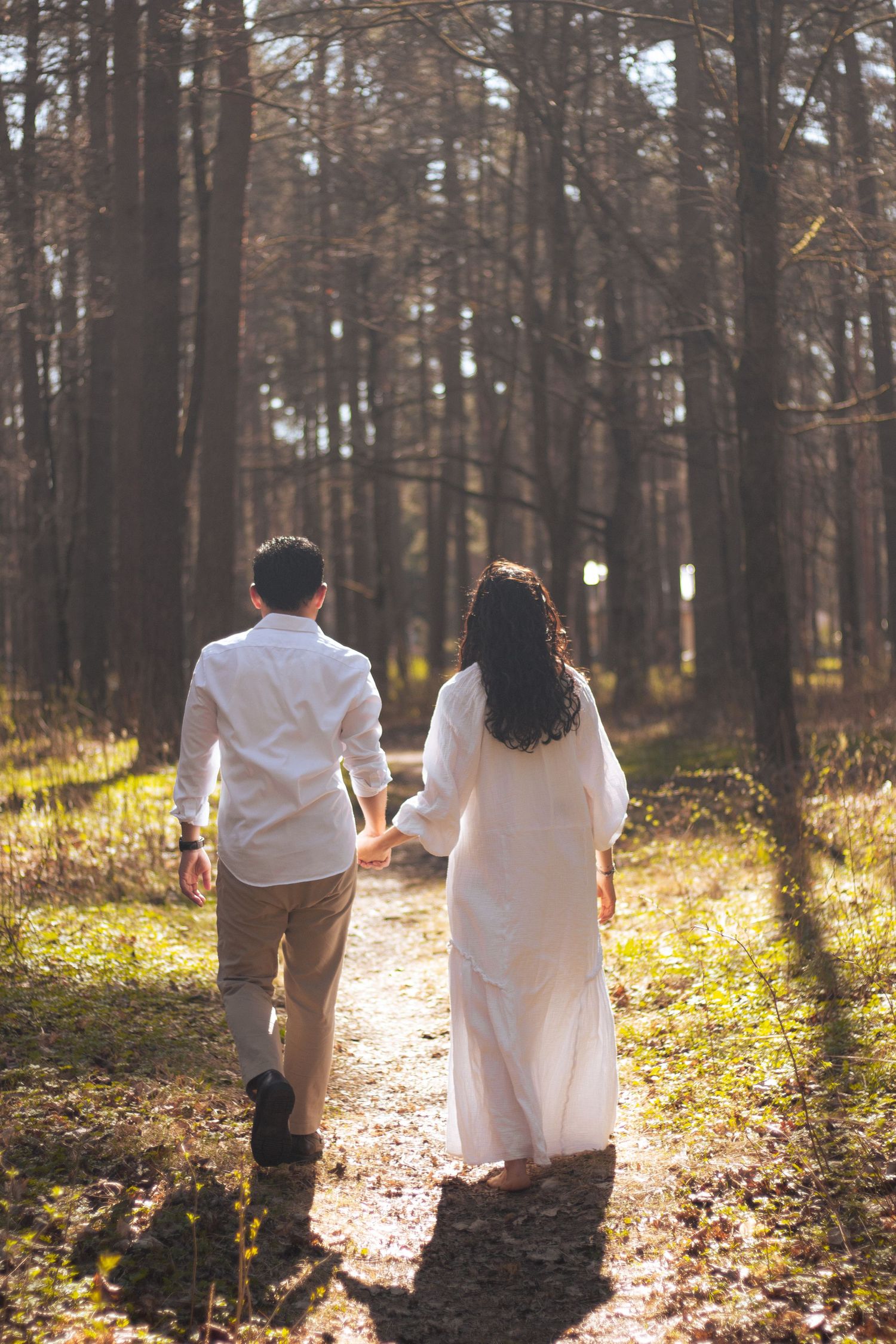 Couple holding hands walking along sunlit forest path, woman in white dress and man in white shirt.