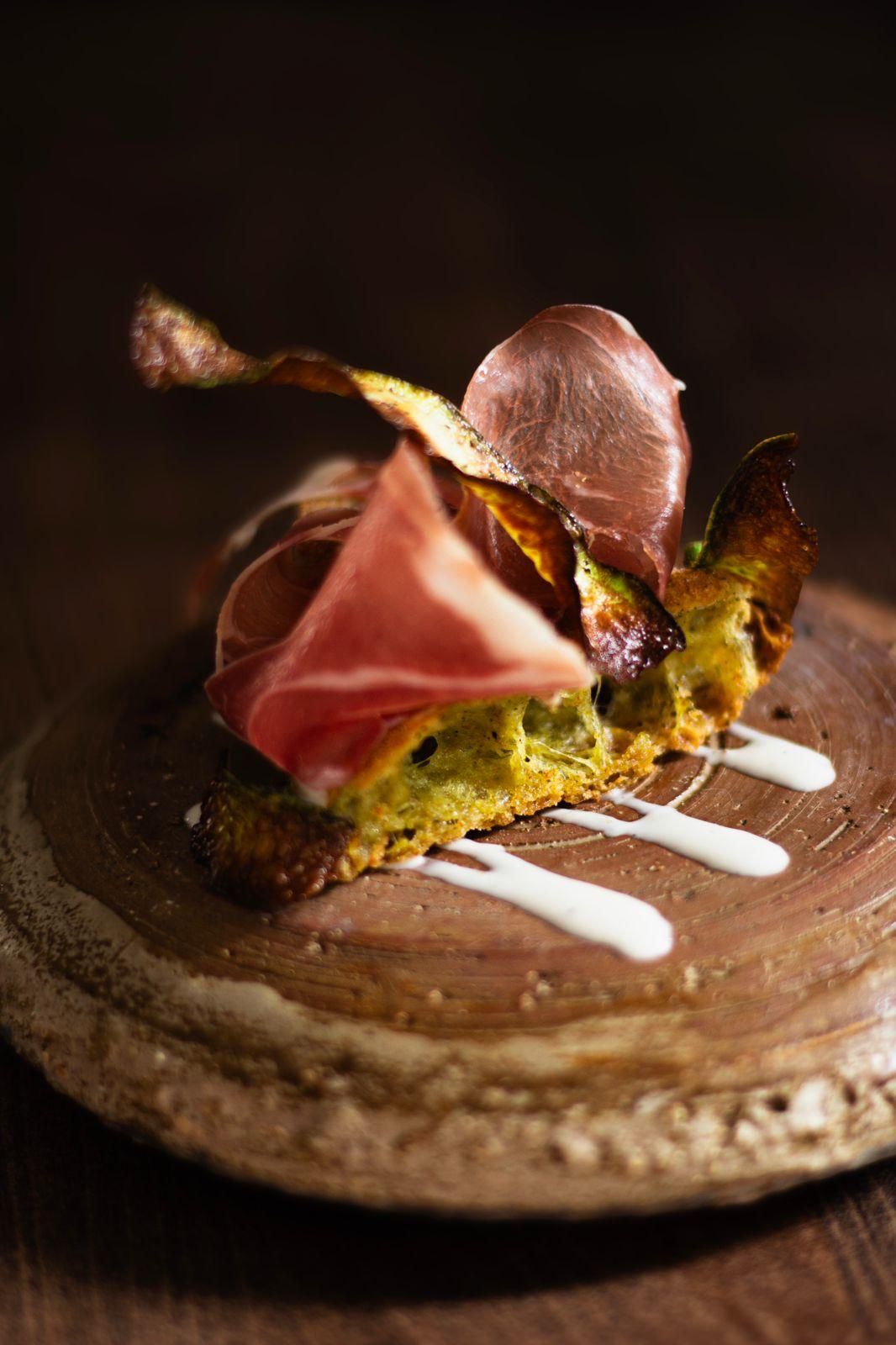 Gourmet open-faced sandwich with cured meat on toasted herb bread, served on rustic wooden board with white sauce.
