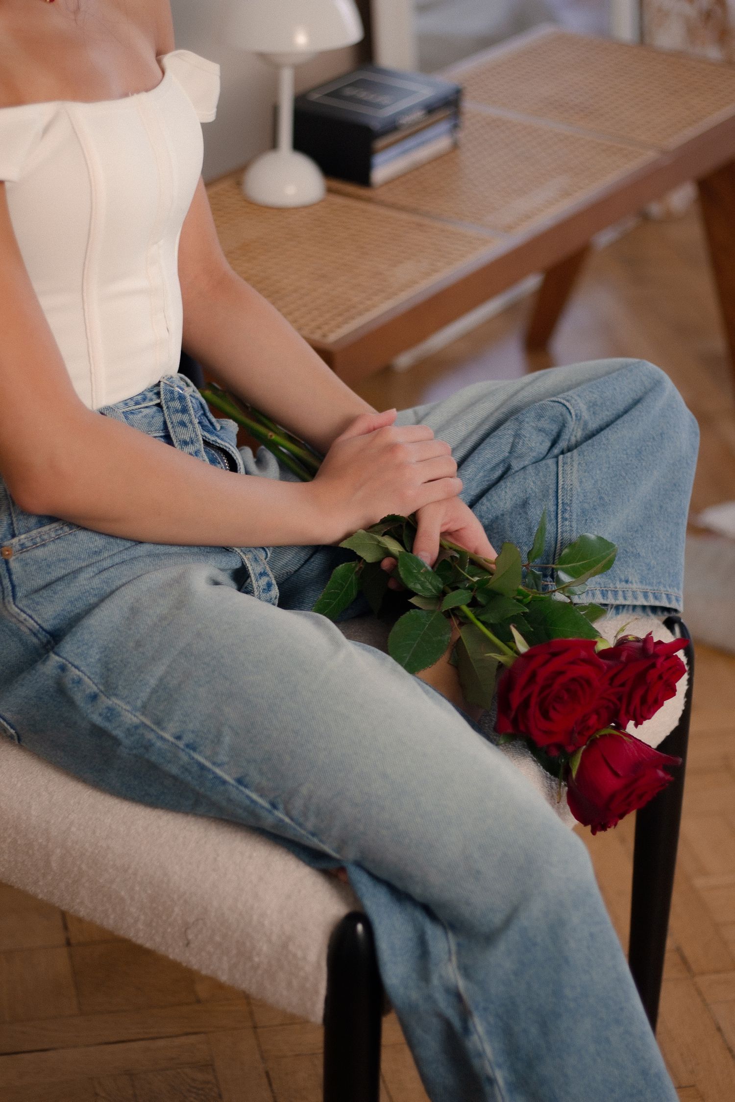 Woman in white top and jeans sitting on chair holding red roses in warmly lit room.
