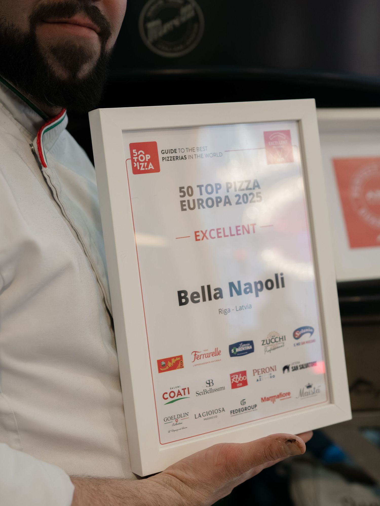 Pizza chef holding a framed 50 Top Pizza Europa 2025 Excellent award certificate for Bella Napoli, Riga.