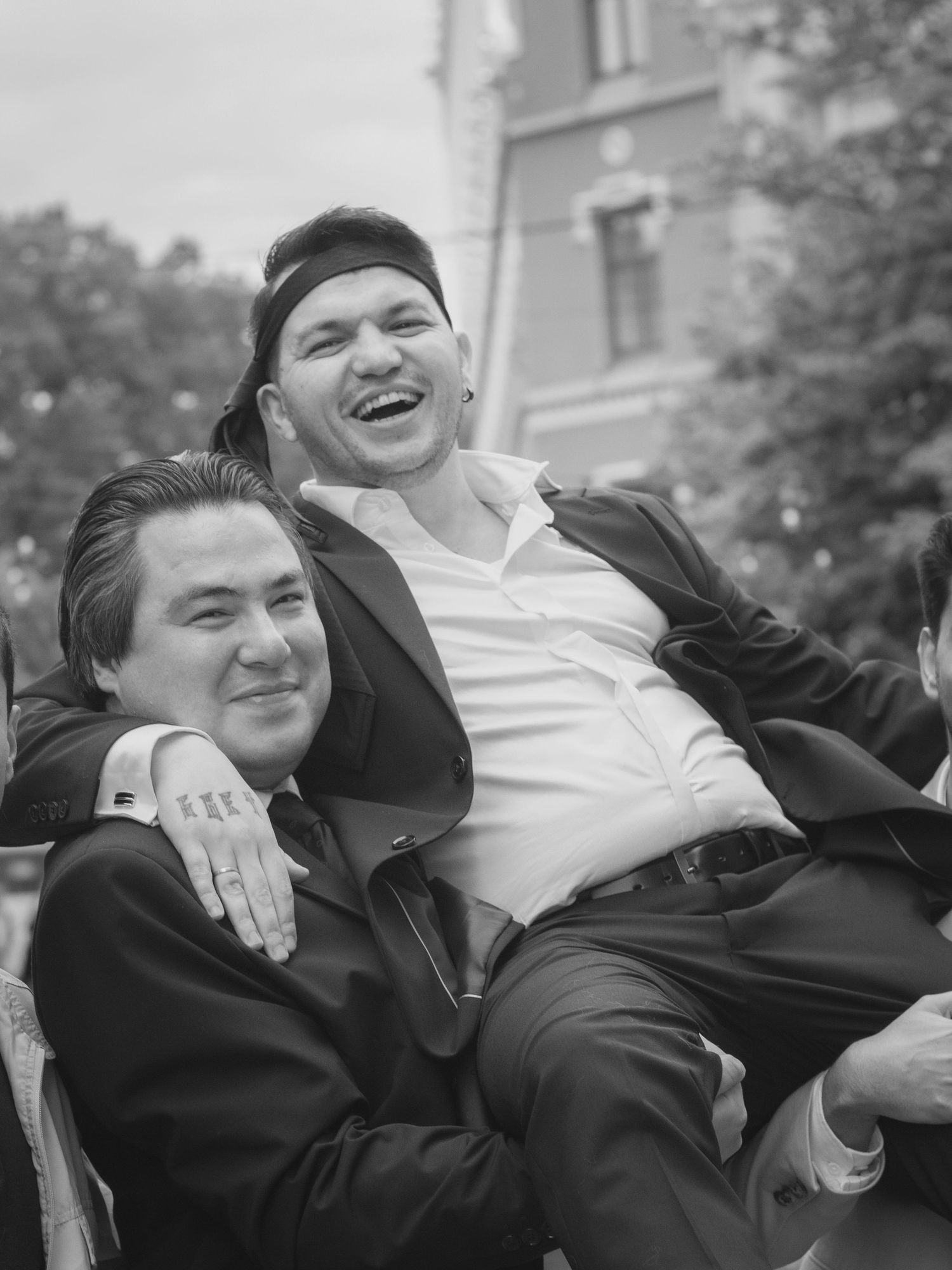 Two men laughing and embracing outdoors in black and white photo, one wearing a backwards cap and white shirt.