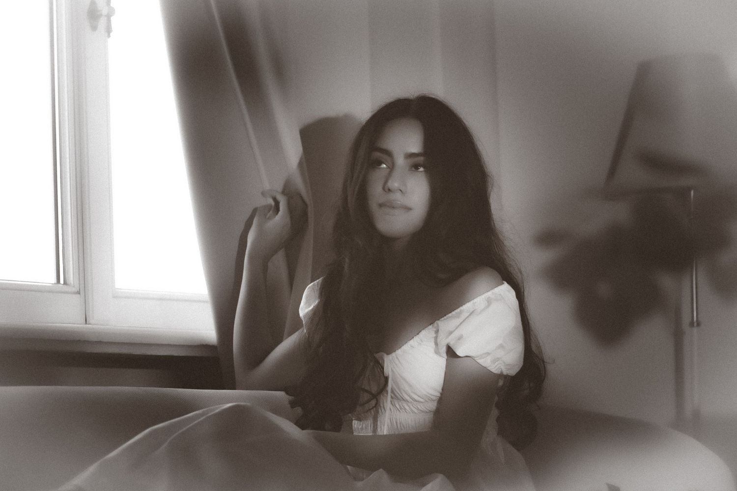 Young woman with long dark hair sitting by window in white dress, vintage black and white photo.