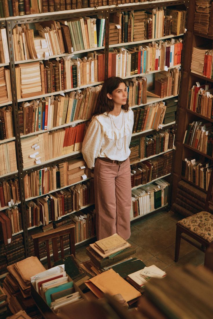 Young woman in white blouse and pink trousers standing among tall bookshelves in a vintage library.