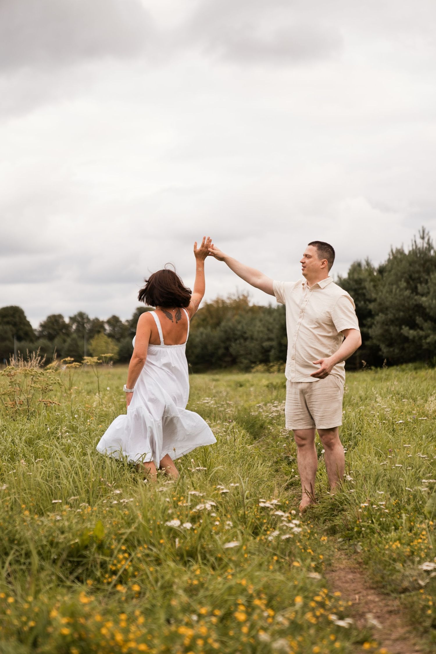 Couple dancing in a wildflower meadow, woman in white dress being twirled by man in beige outfit under cloudy sky.