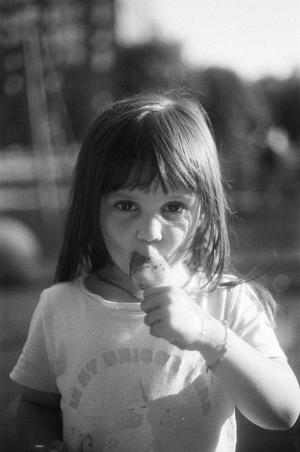 Black and white photo of a young girl with straight hair eating an ice cream cone outdoors.
