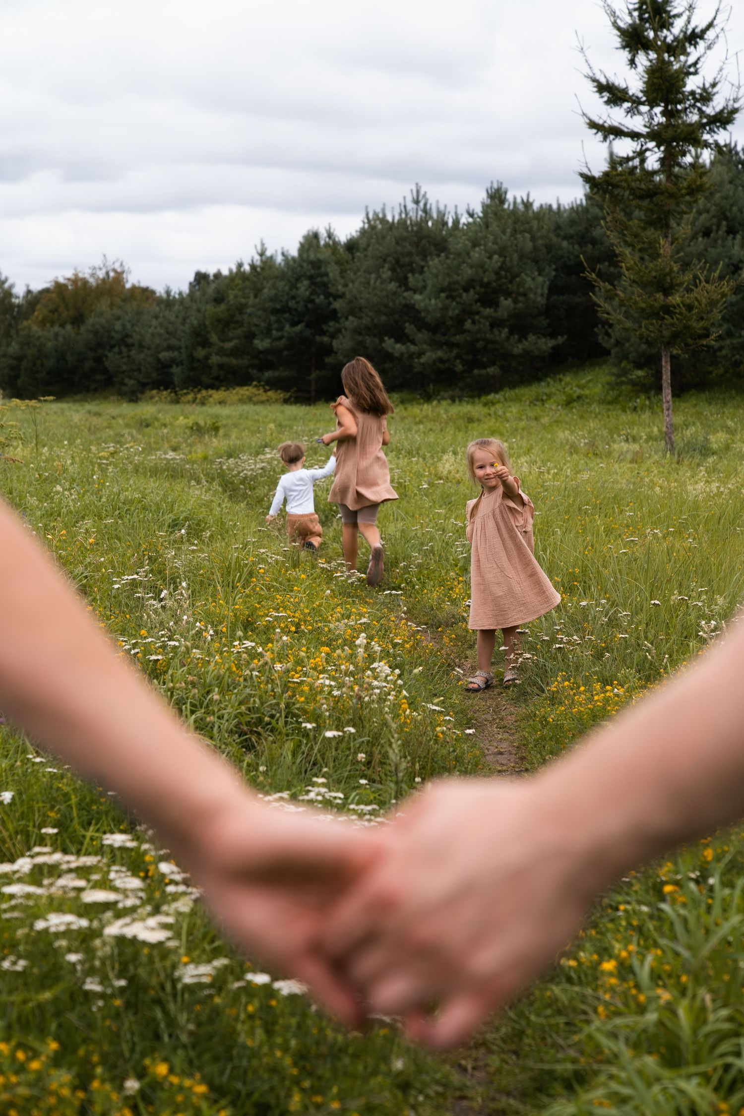 Adults hold hands while three children run through a wildflower meadow near a forest on a cloudy day.