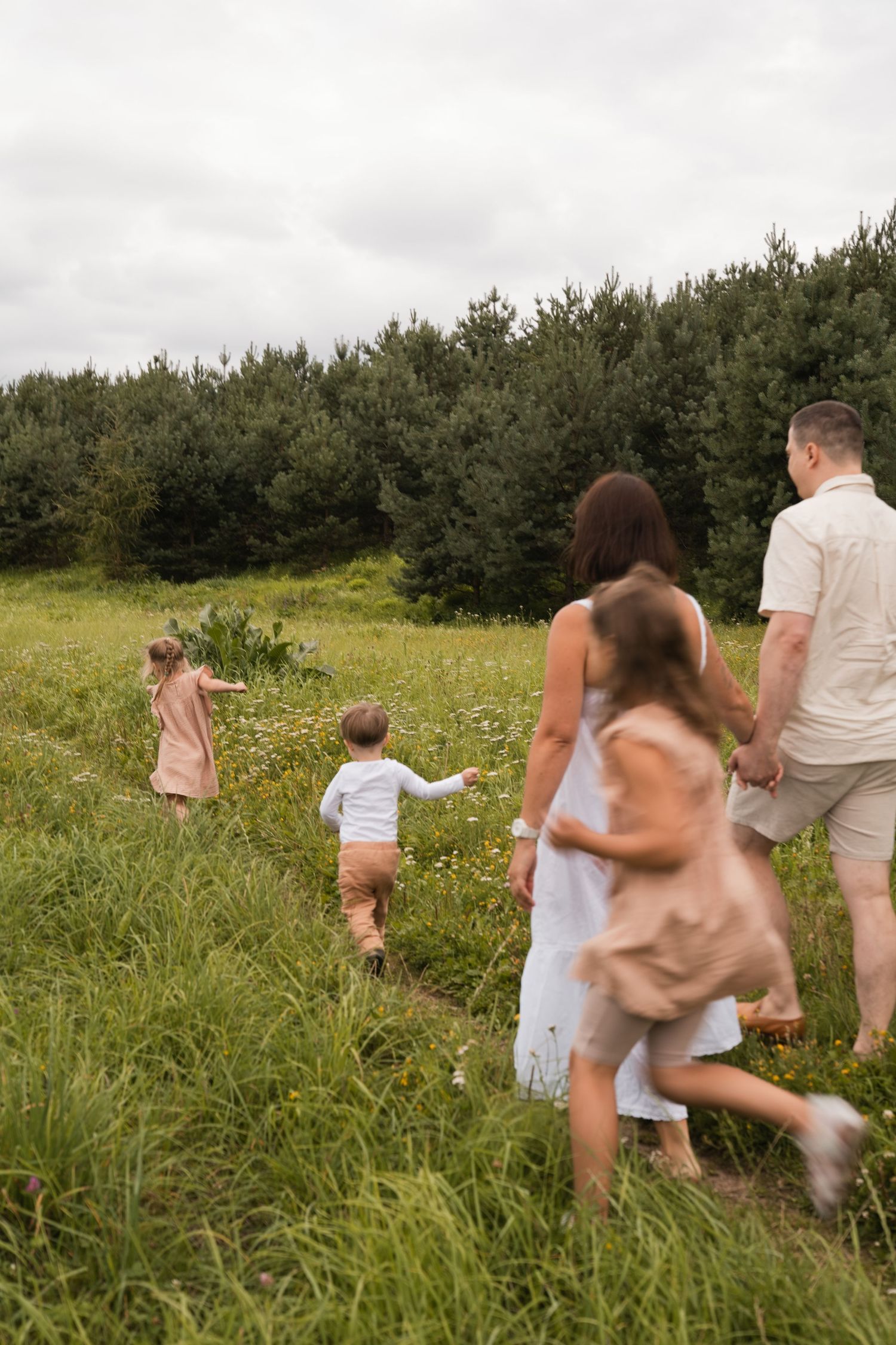 Family walking through a lush green meadow toward a forest on a cloudy day.