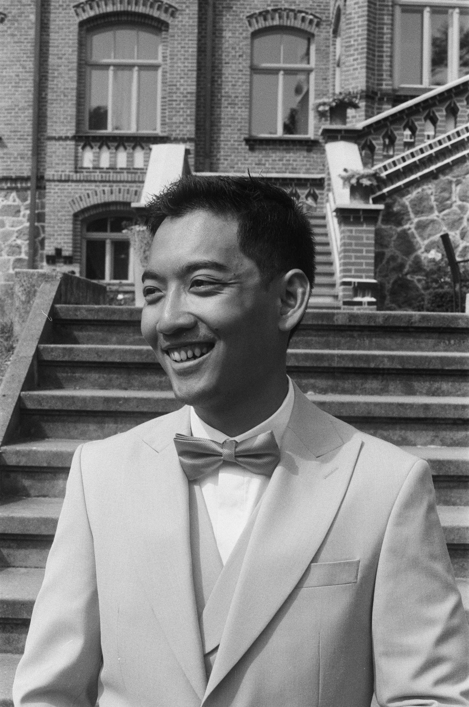 Smiling groom in white suit and bow tie standing before historic brick building.