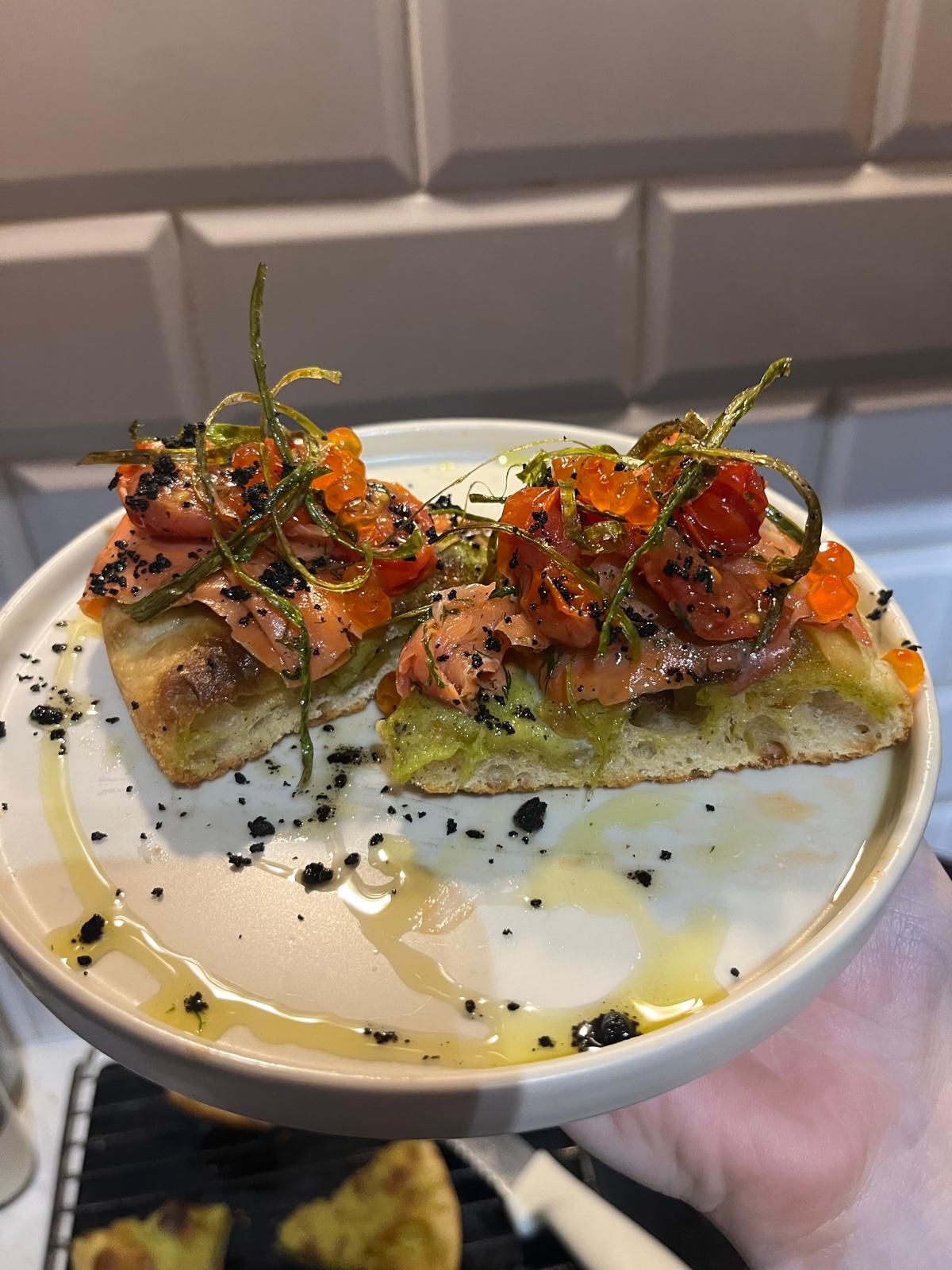 Artisan focaccia topped with smoked salmon, cherry tomatoes, black sesame seeds, and herb oil on white plate.